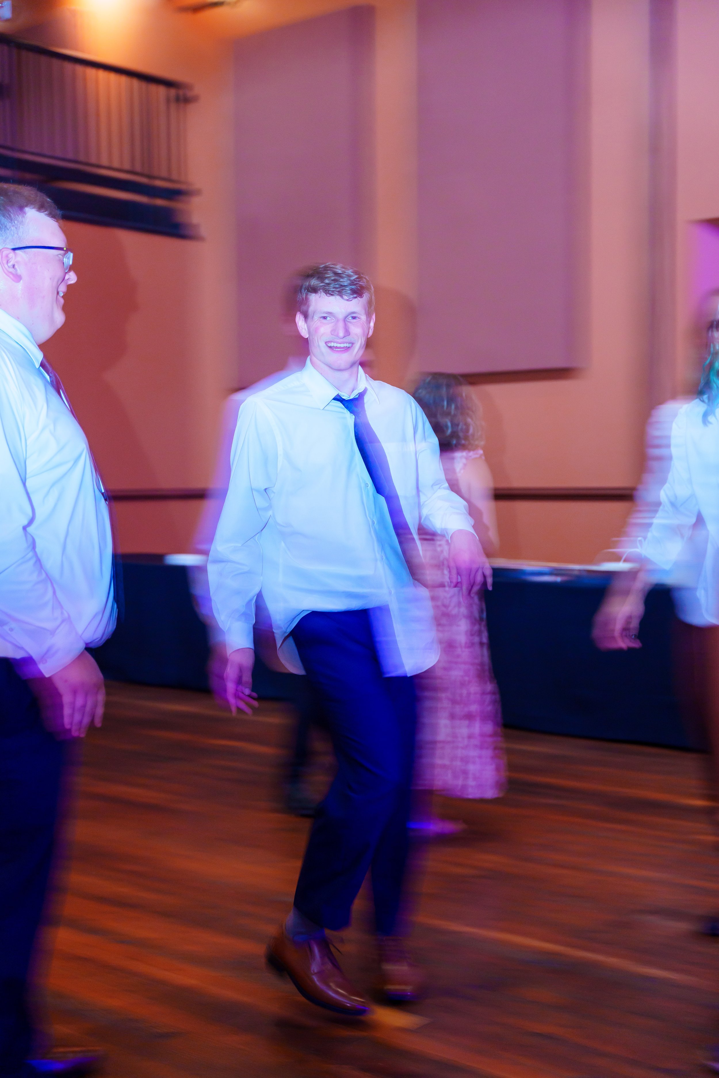 Young man with curly hair, white shirt, and blue tie, smiling and walking, surrounded by other people dancing or socializing in a dimly lit room with purple and orange lighting and wooden floors.