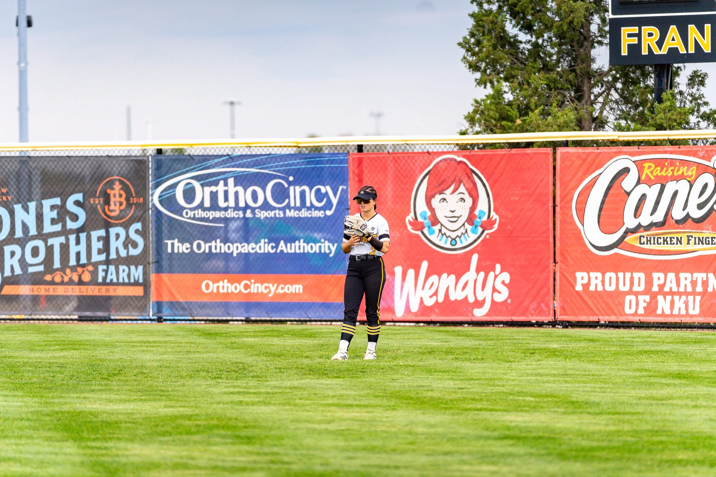 A female baseball player standing on a field wearing a uniform and cap, holding a glove, with advertising banners behind her on a fence.