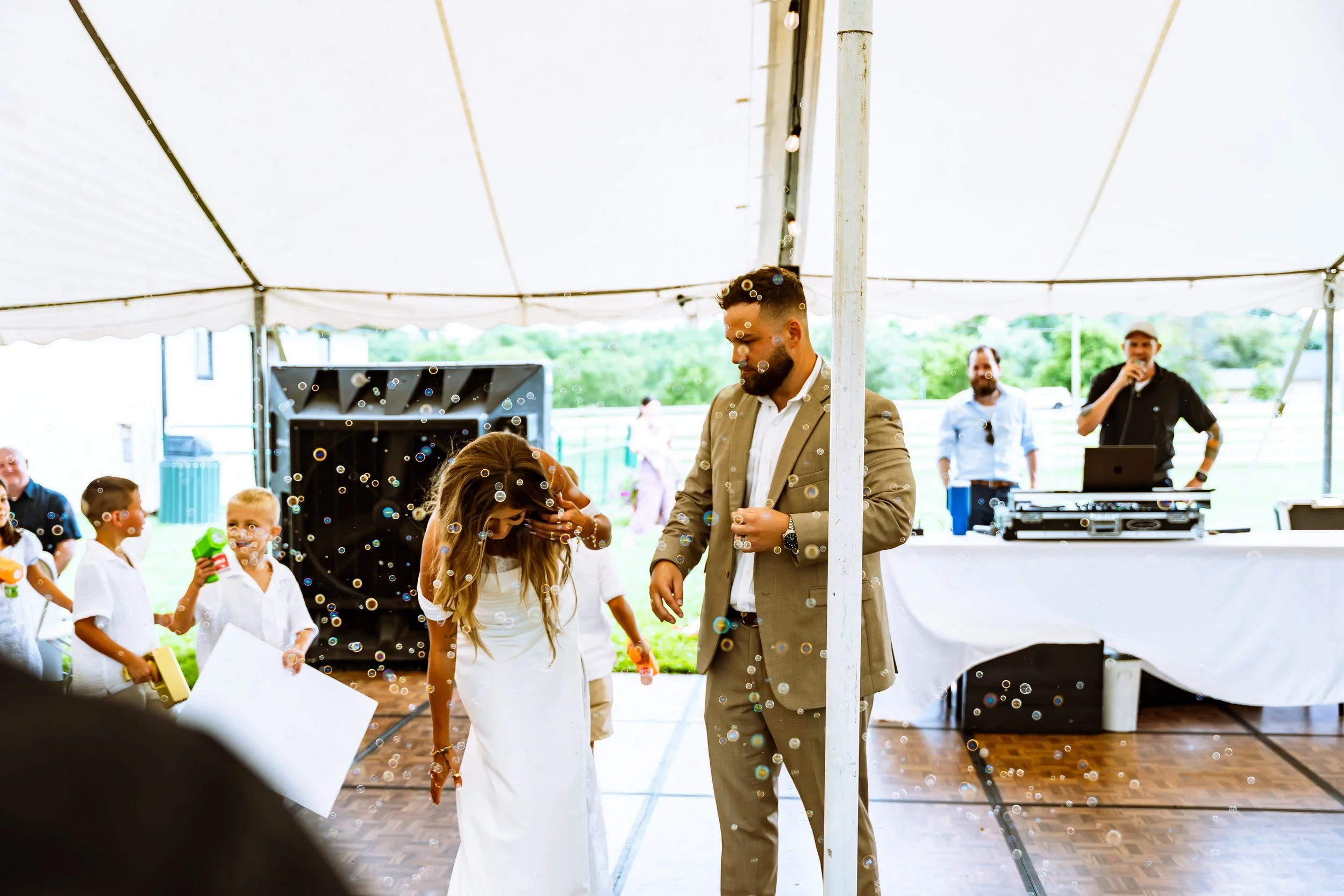 A man in a tan suit and a woman with long hair in a white dress are dancing, surrounded by children and adults under a large white tent at an outdoor event. Bubbles float in the air, and there is a DJ setup with a speaker and mixing equipment in the 