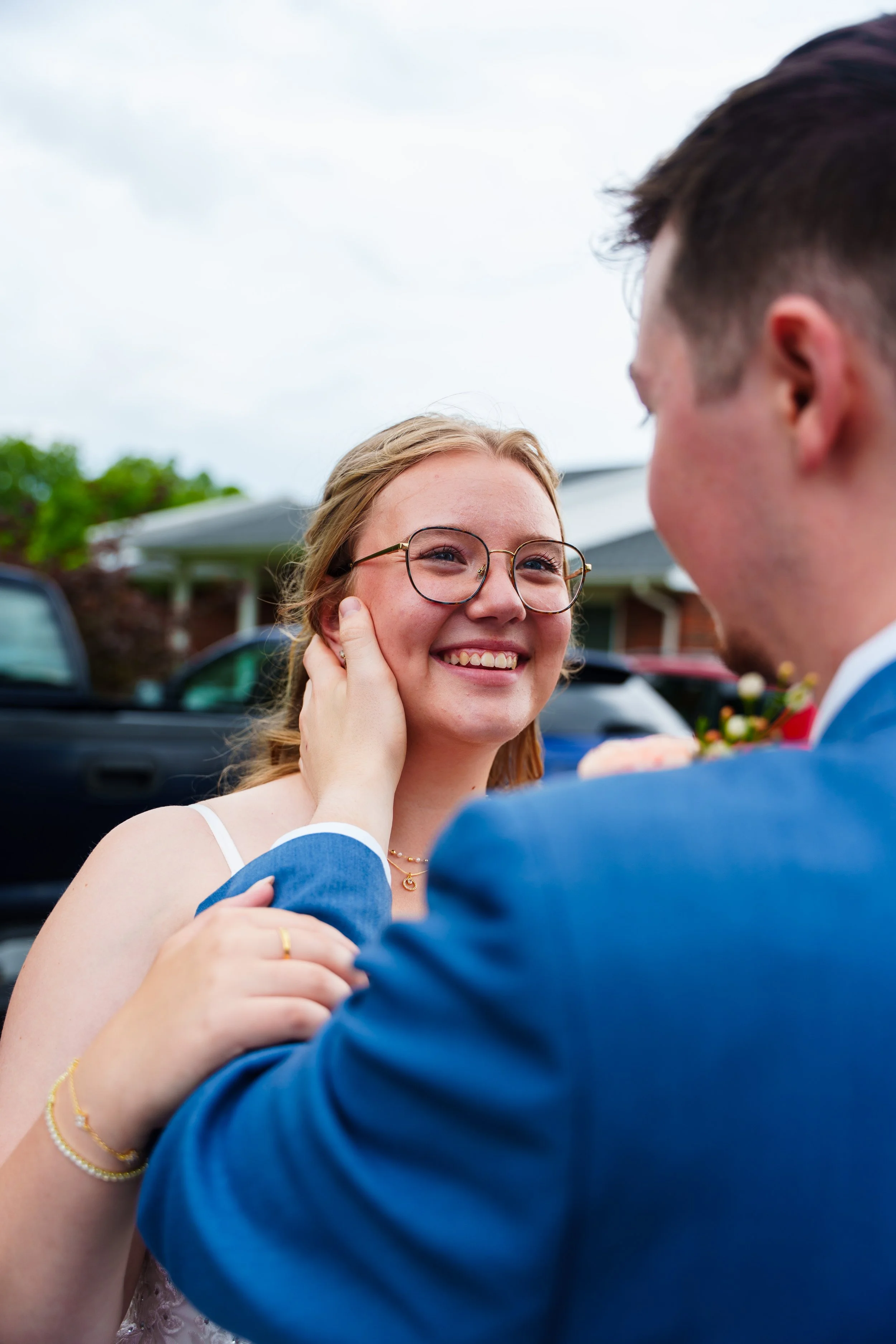 A smiling woman with glasses and a white dress is being touched on the face by a person in a blue suit, with an outdoor setting and other cars in the background.