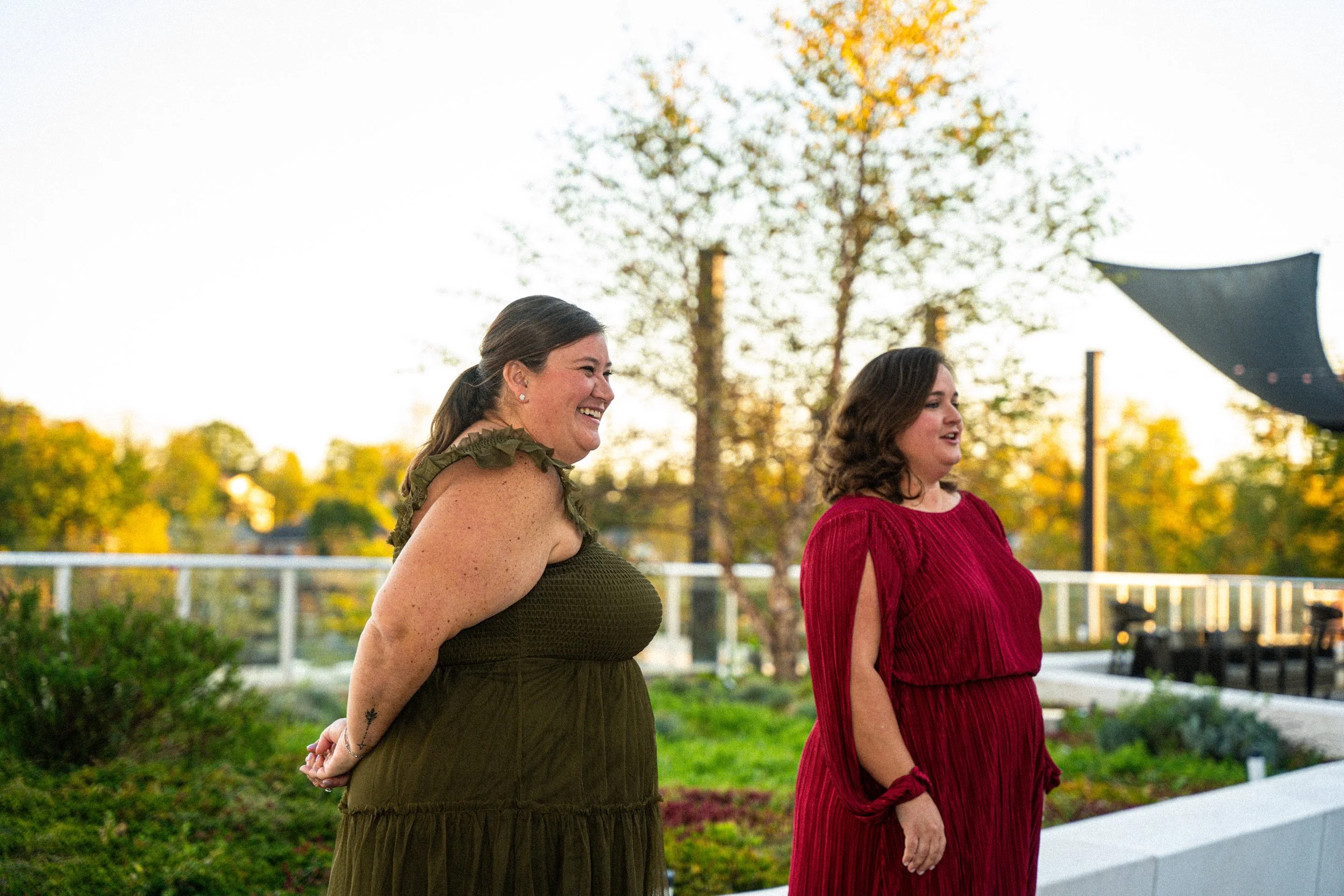 Two women standing outdoors on a grassy area in the evening, looking to the right and smiling. One woman has brown hair and is wearing a red dress; the other woman has dark hair and is wearing an olive-green dress. There are trees with yellow leaves 