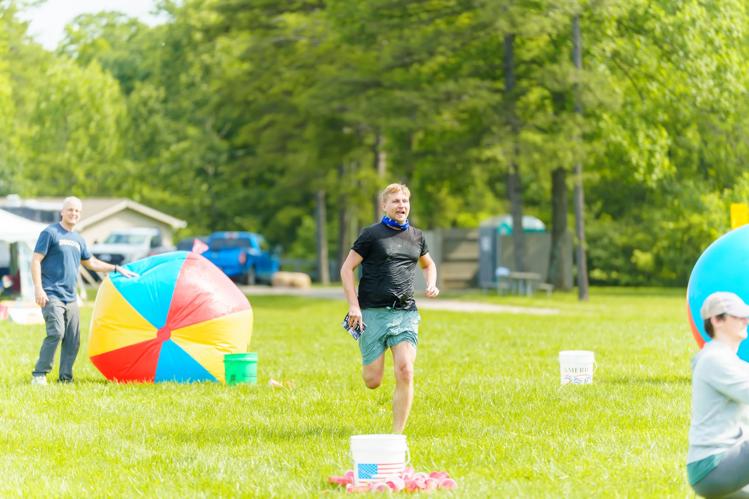 A young man in shorts and a black T-shirt playing a game at an outdoor event with large beach balls and various equipment on a grassy field, with people and trees in the background on a sunny day.