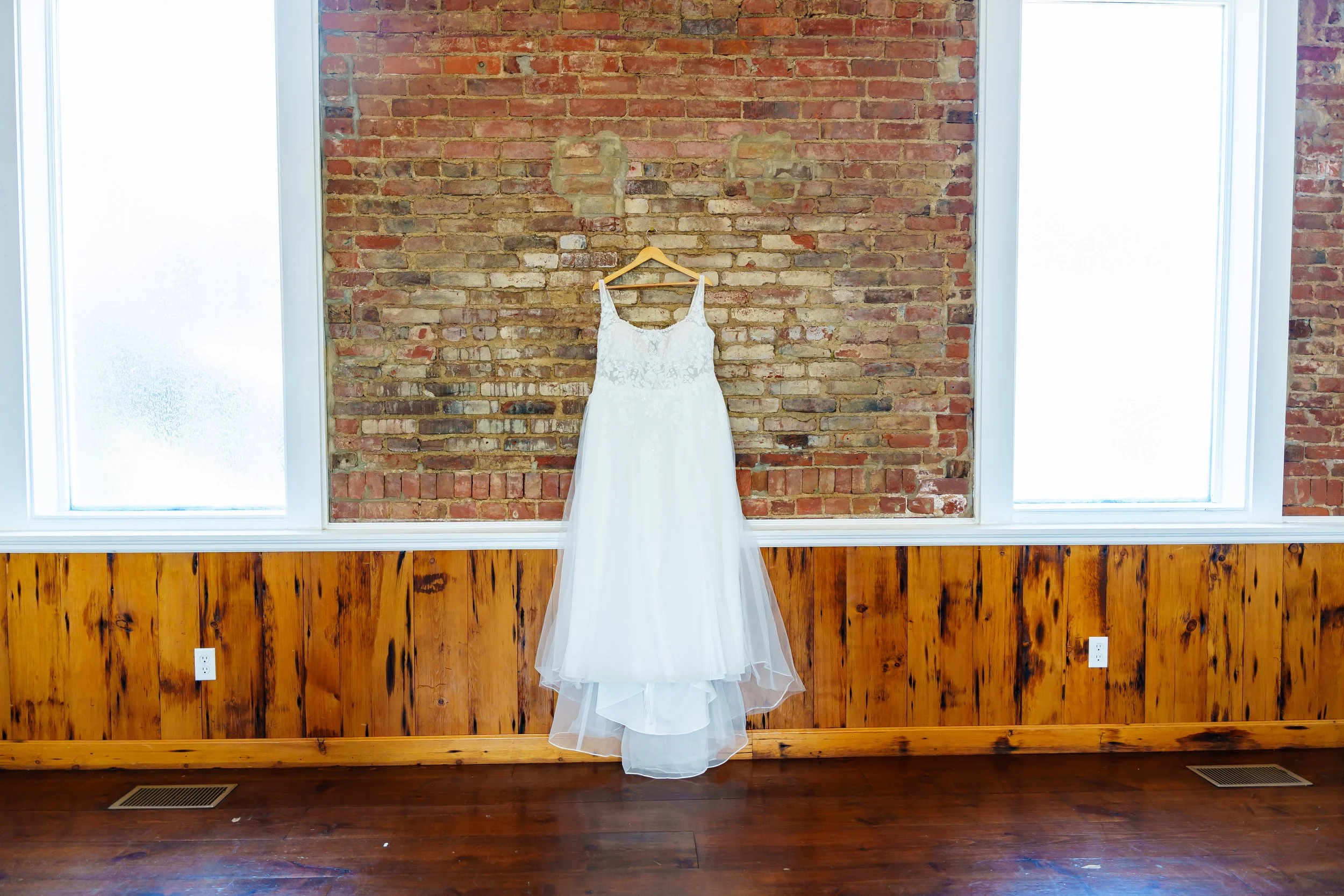 A white wedding dress hanging on a wooden hanger against a brick wall, with two large windows on either side providing natural light.