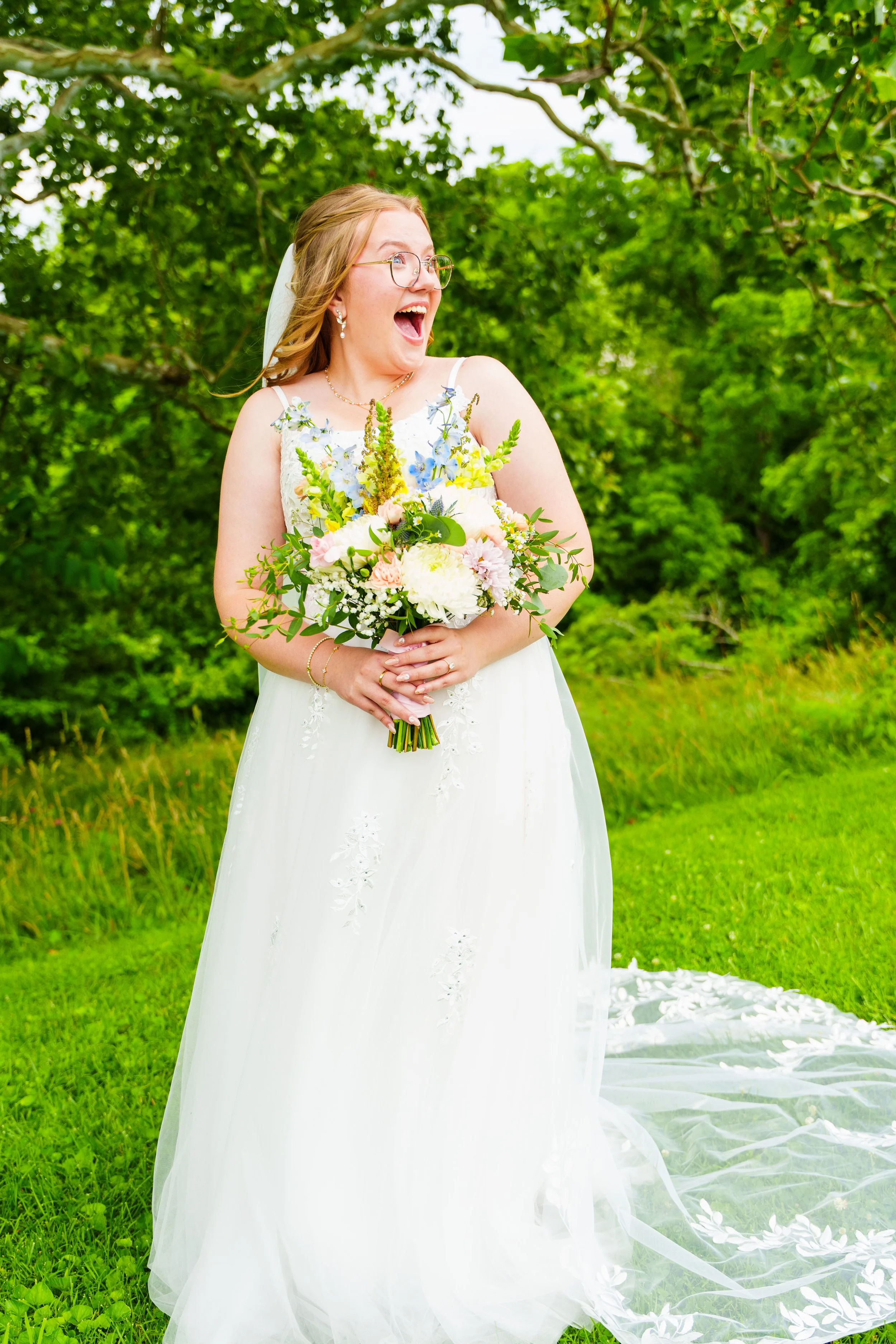 A joyful woman in a white wedding dress holding a bouquet of flowers outdoors, surrounded by green trees and grass.