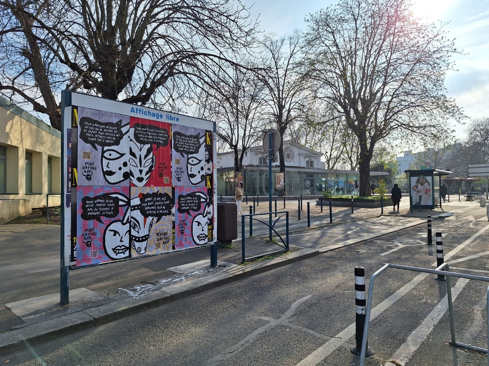 City street scene with a free poster display board featuring black and white illustrations and French text, trees without leaves, and a modern bus stop shelter in the background, with a few pedestrians walking.