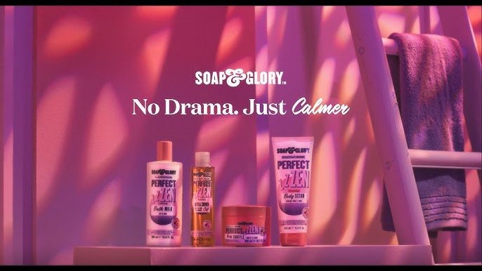 Display of Soap & Glory skincare products on a pink shelf with a purple towel on a ladder in the background, with a pink and purple lighting ambiance. The text reads 'Soap & Glory: No Drama. Just Calmer'.