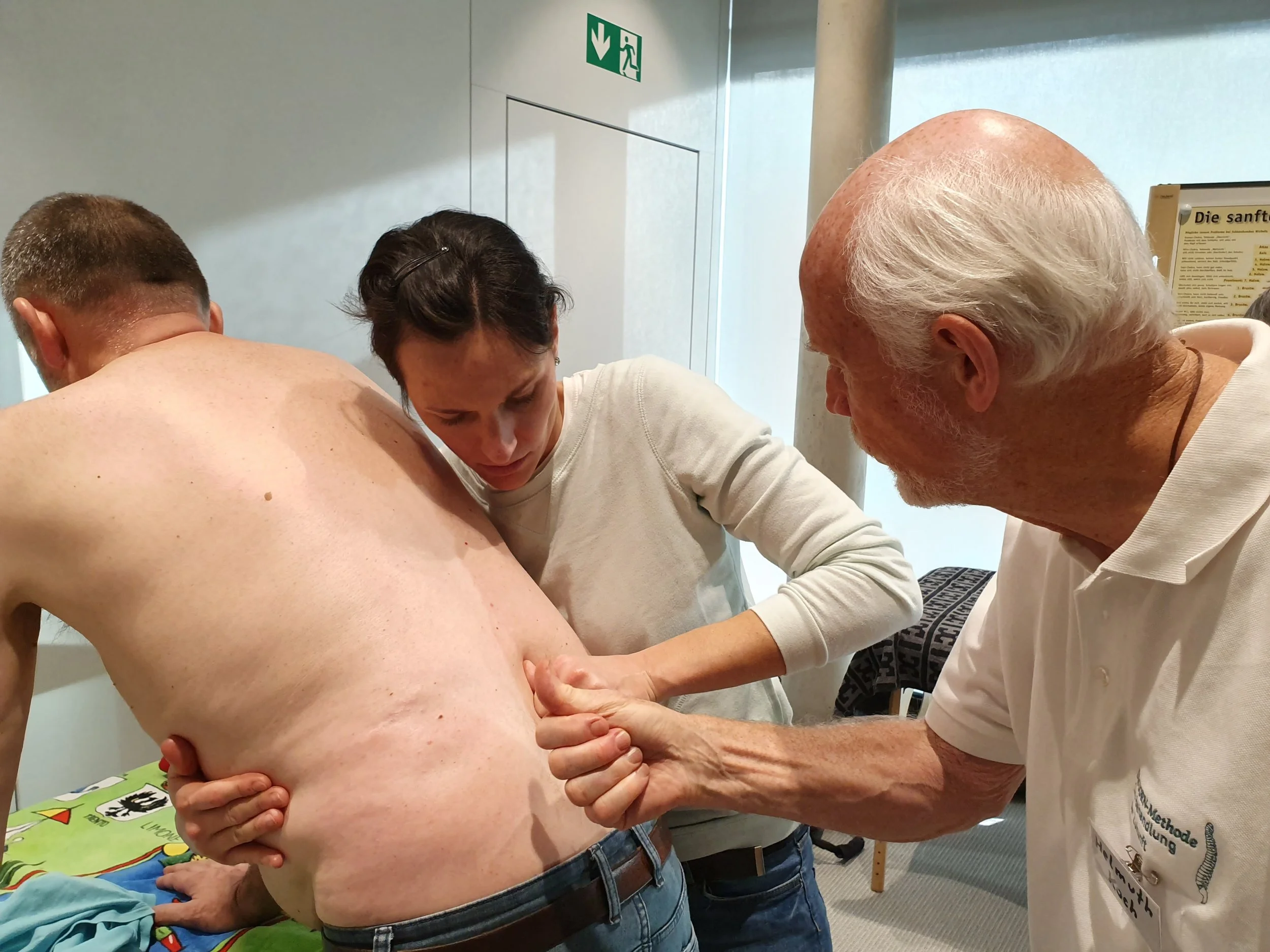 Drei Personen sind in einem Seminarraum an einer Behandlung beteiligt. Ein Mann mit freiem Oberkörper und dunklen Jeans, dessen Rücken zum Betrachter gewandt ist, wird von einer Frau in einem hellen Pullover und Jeans behandelt.