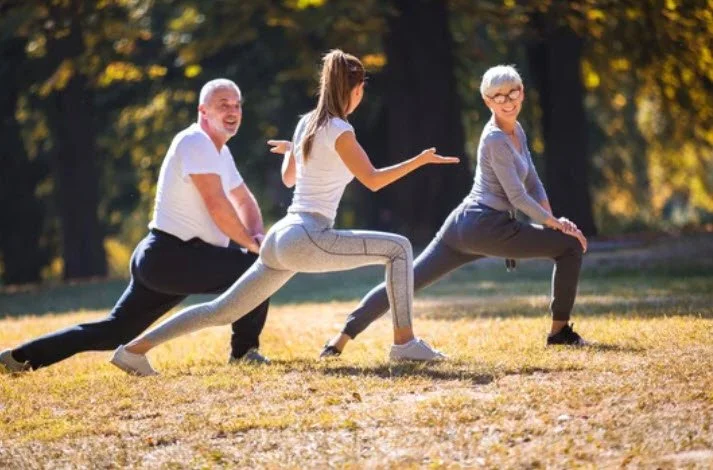 Ein Mann und eine Frau machen im Freien sportliche Dehnübungen unter Anleitung einer jungen sportlichen Frau. Diese hat ihr rechtes Bein nach vorne gestreckt, während sie in der Hocke ist. Sie trägt ein weißes T-Shirt.