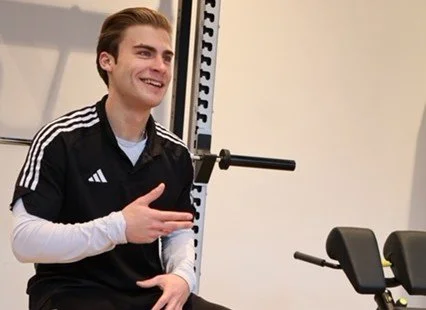 Ein lächelnder, gestikulierender junger Mann mit dunklen Haaren sitzt in einem Raum, der wie ein Fitnessstudio aussieht. Er trägt ein schwarzes Poloshirt mit weißen Streifen an den Ärmeln und ein weißes Langarmshirt darunter.