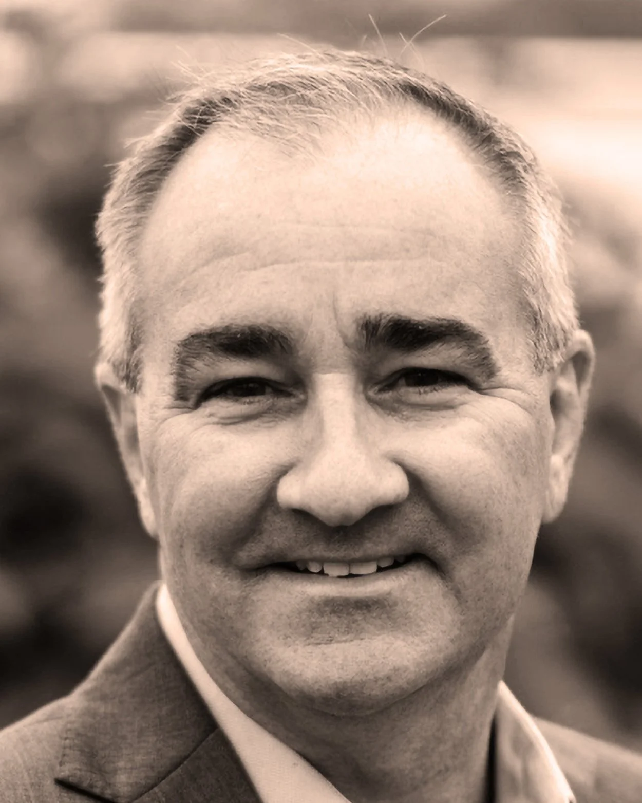 Close-up black and white photo of a middle-aged man smiling, with short hair, wearing a suit and collared shirt.