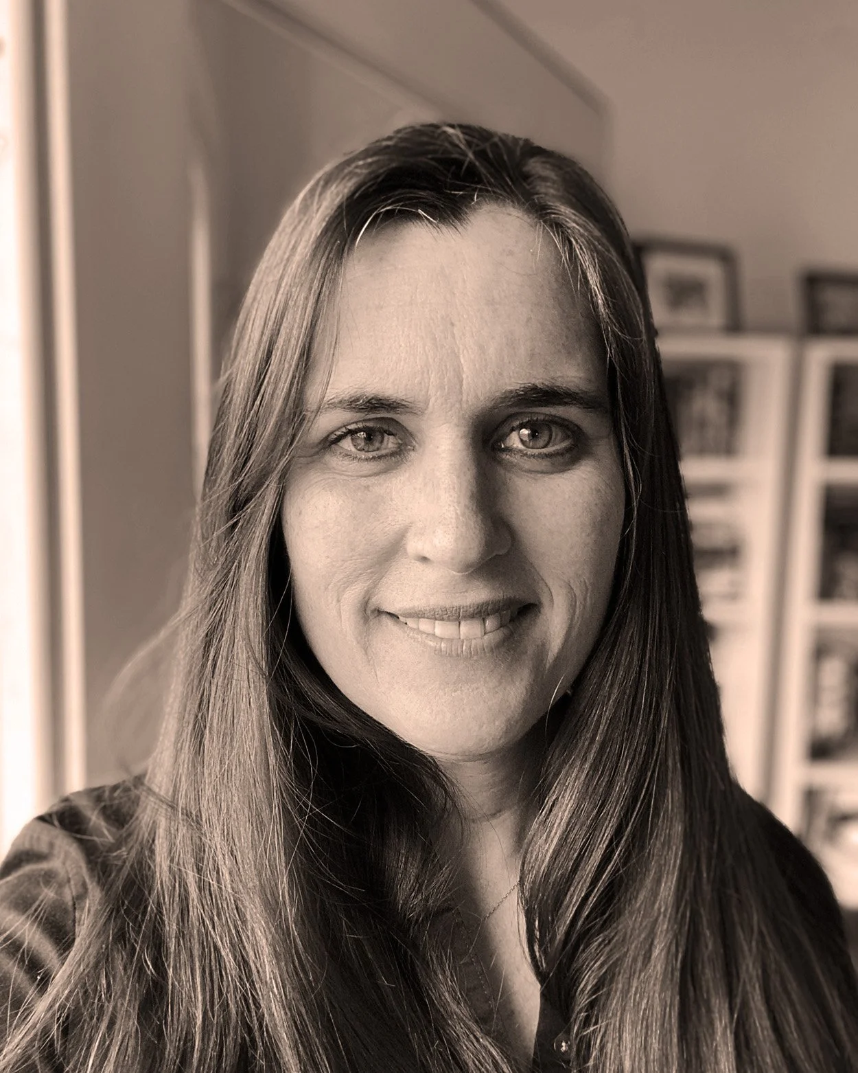 A black-and-white portrait of a woman with long hair, smiling, indoors with shelves and framed pictures in the background.