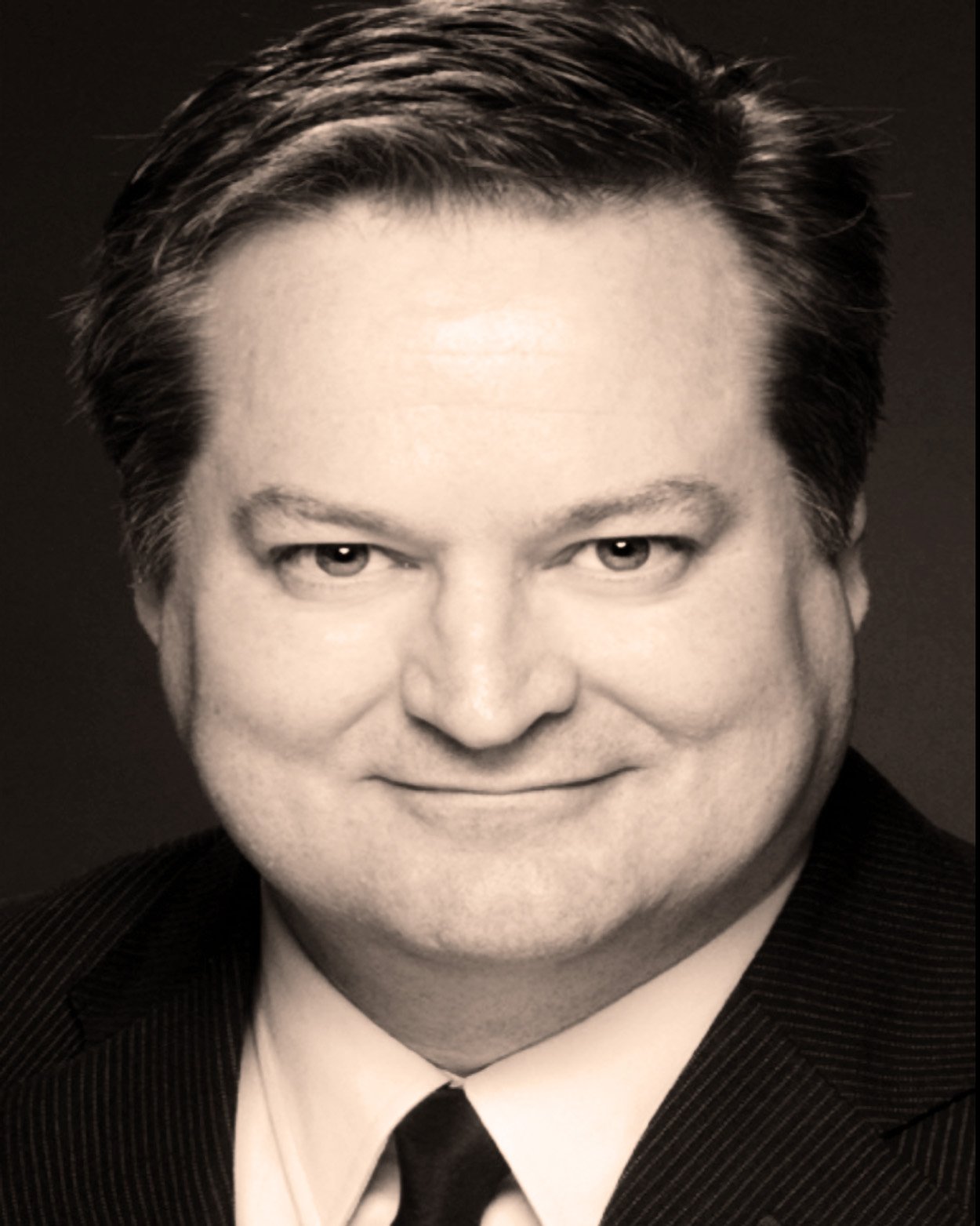 Black and white headshot of a man with dark, styled hair, wearing a suit and tie, smiling slightly.