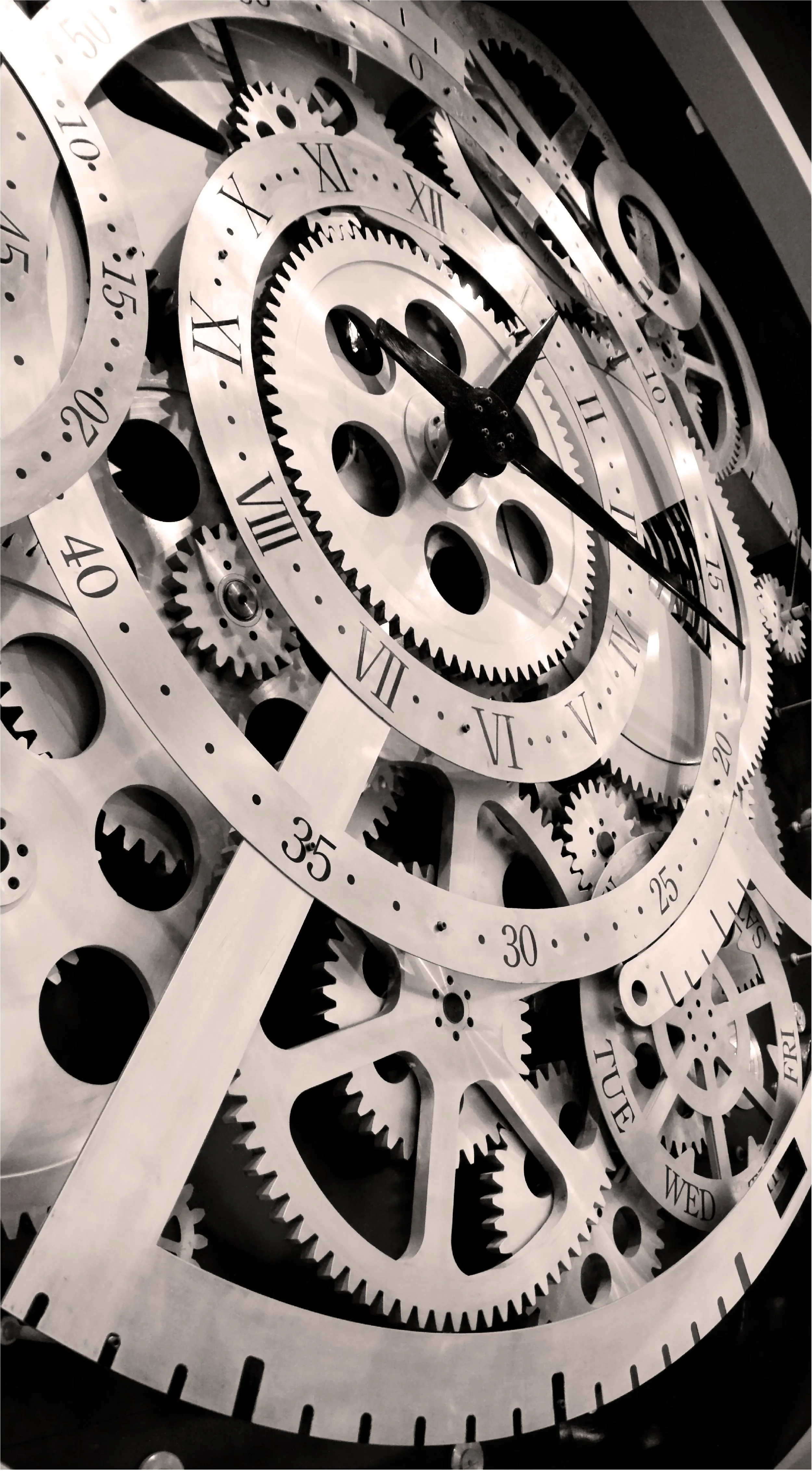 Close-up of a complex mechanical clock or gear system with multiple interconnected gears and watch dials, some with Roman numerals, in black and white.