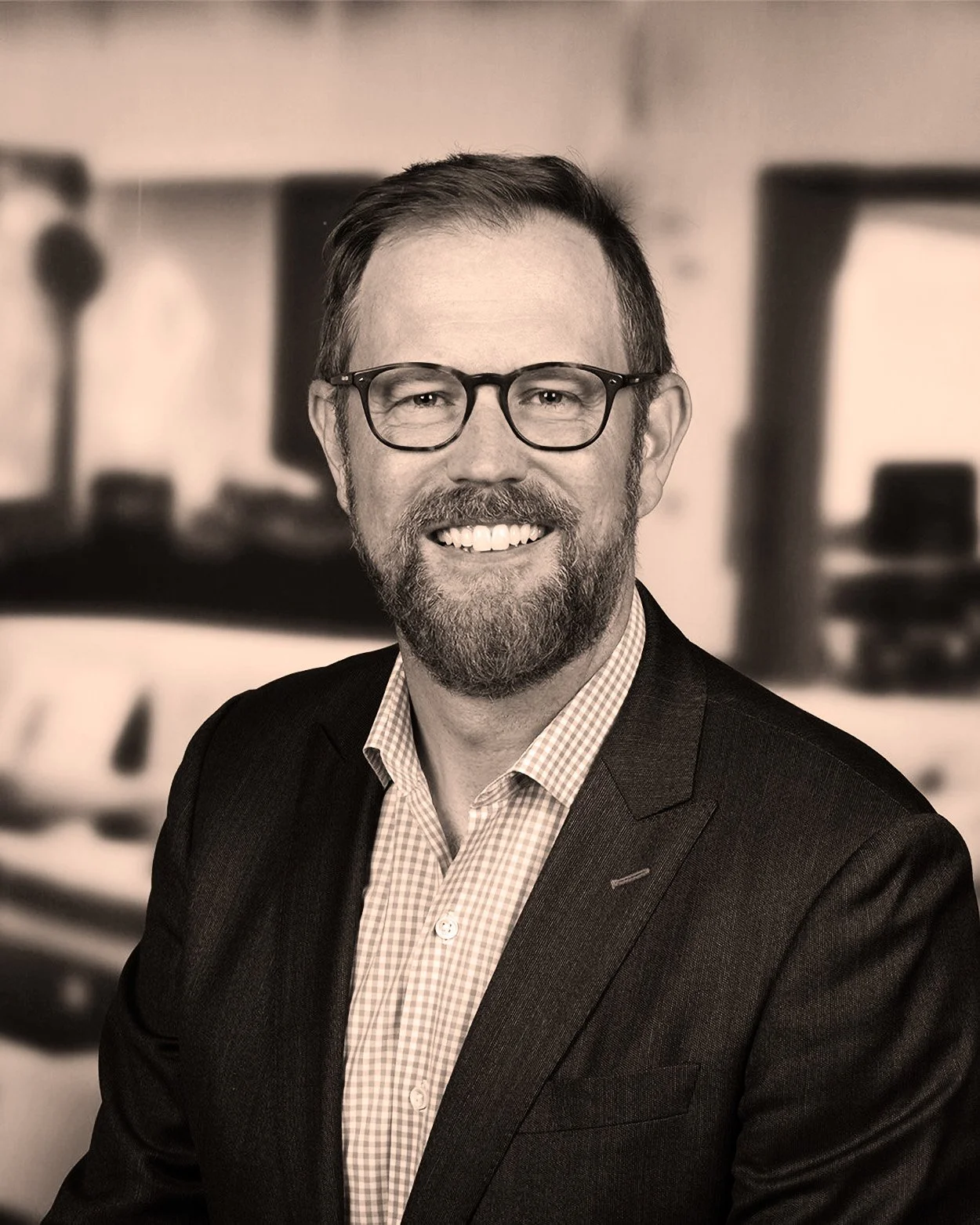 A smiling man with glasses and a beard wearing a suit and checkered shirt, in an office setting.