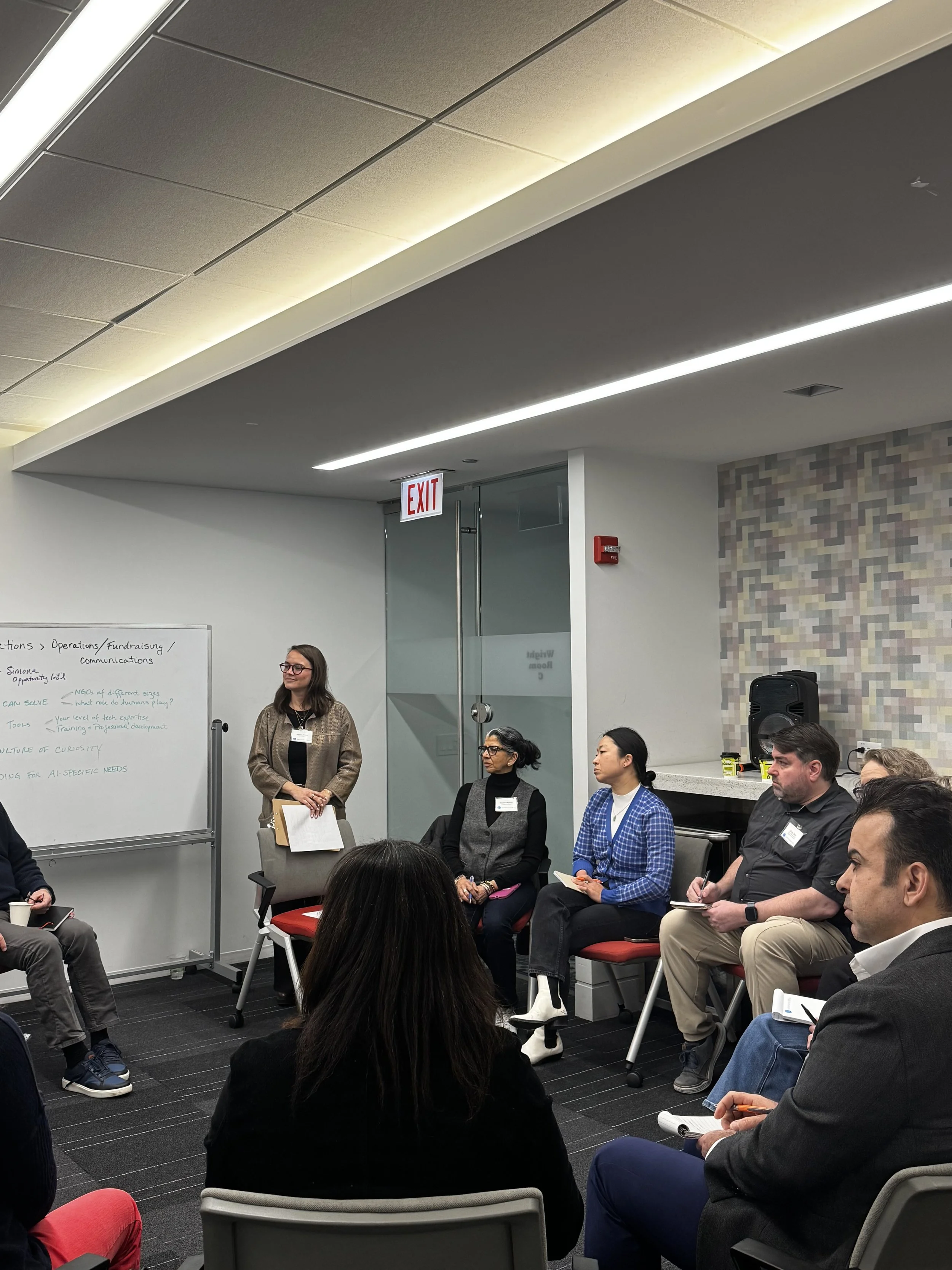 A group of diverse people participating in a discussion or workshop in a modern conference room with a whiteboard, some seated and one woman standing near the whiteboard.