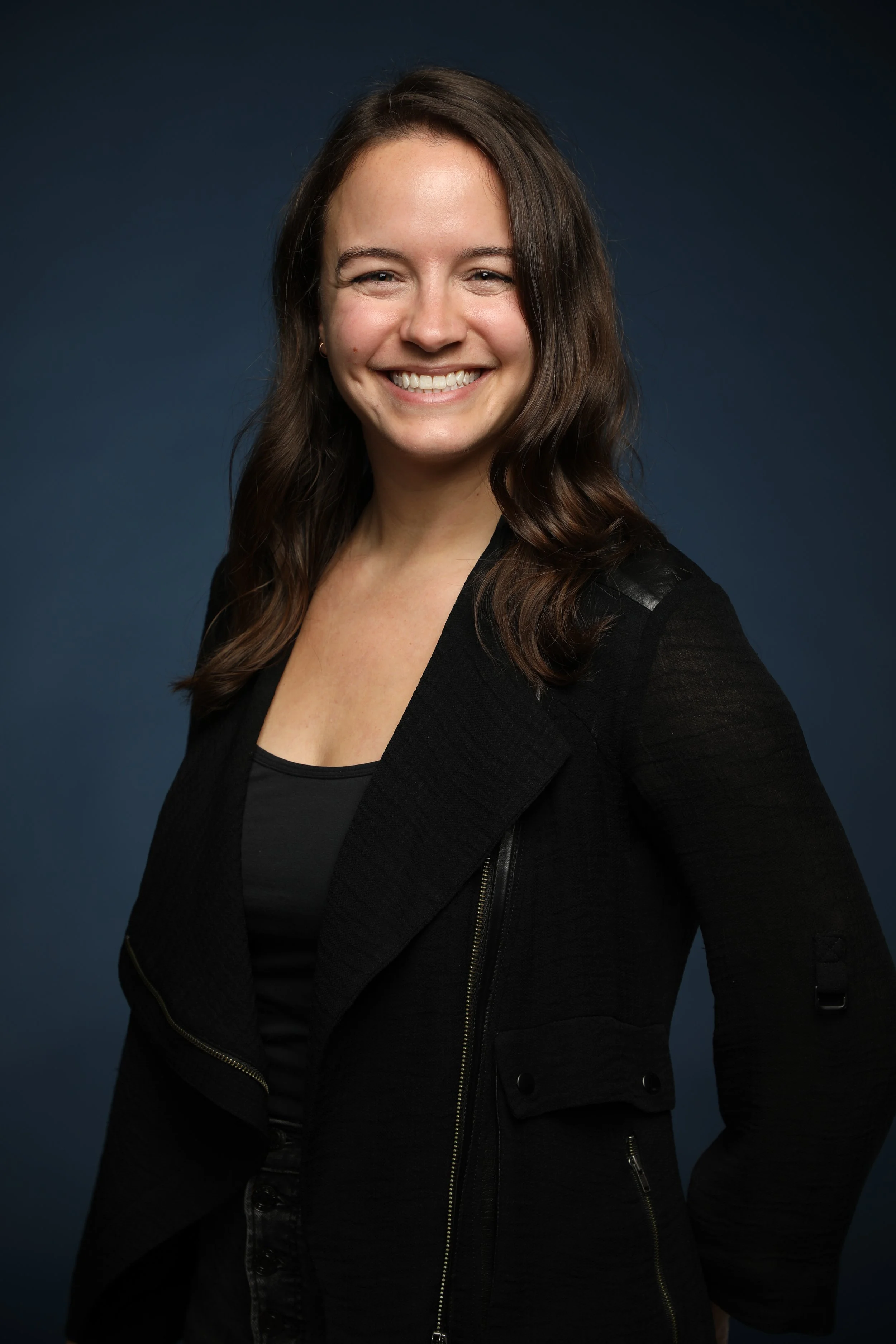 A woman with wavy brown hair, smiling, wearing a black top and black jacket, posing against a dark blue background.