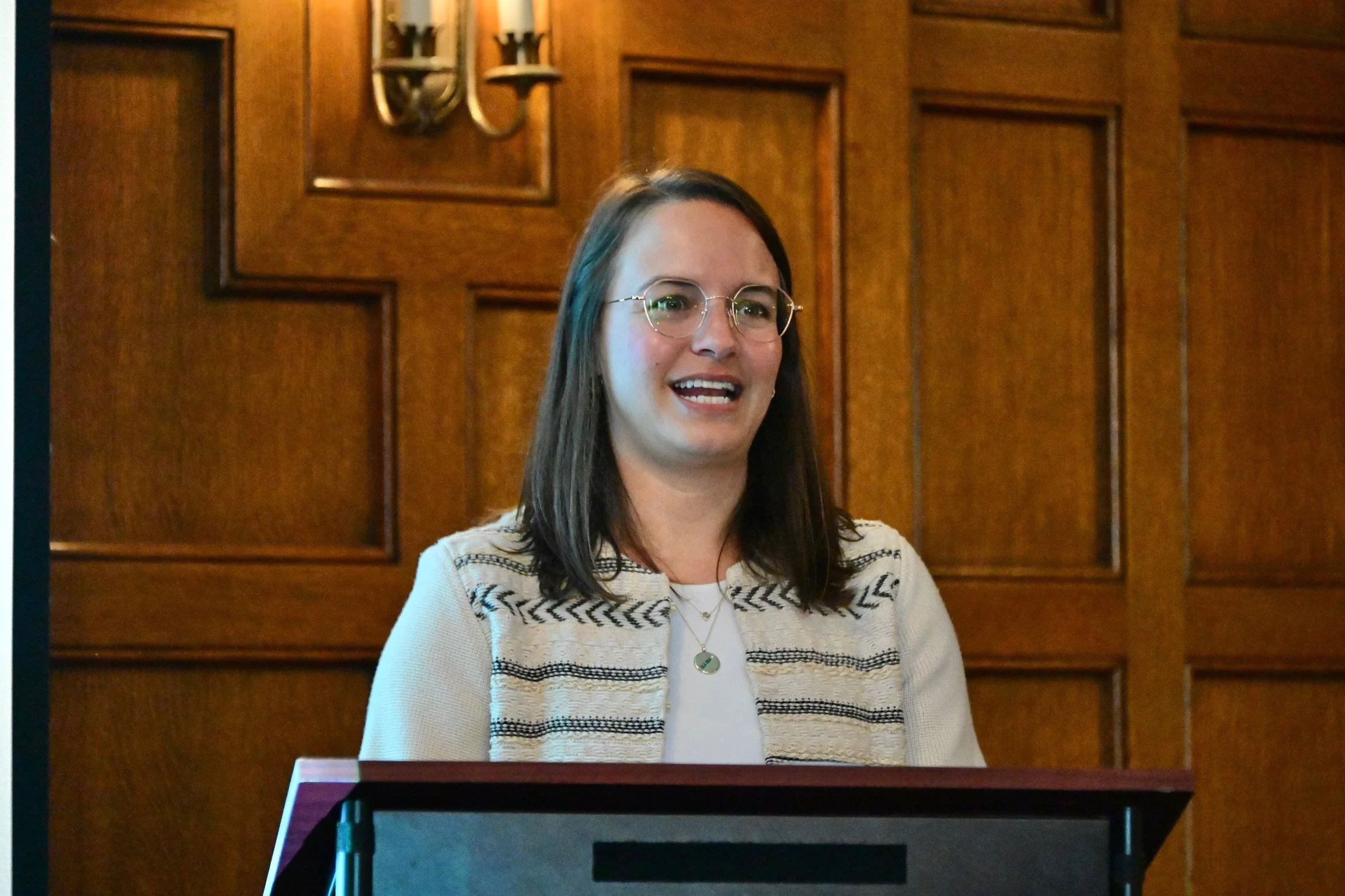 A woman with shoulder-length dark hair, wearing glasses, a cream and black patterned blazer, and a white top, is speaking at a podium with a wooden wall and a wall sconce in the background.
