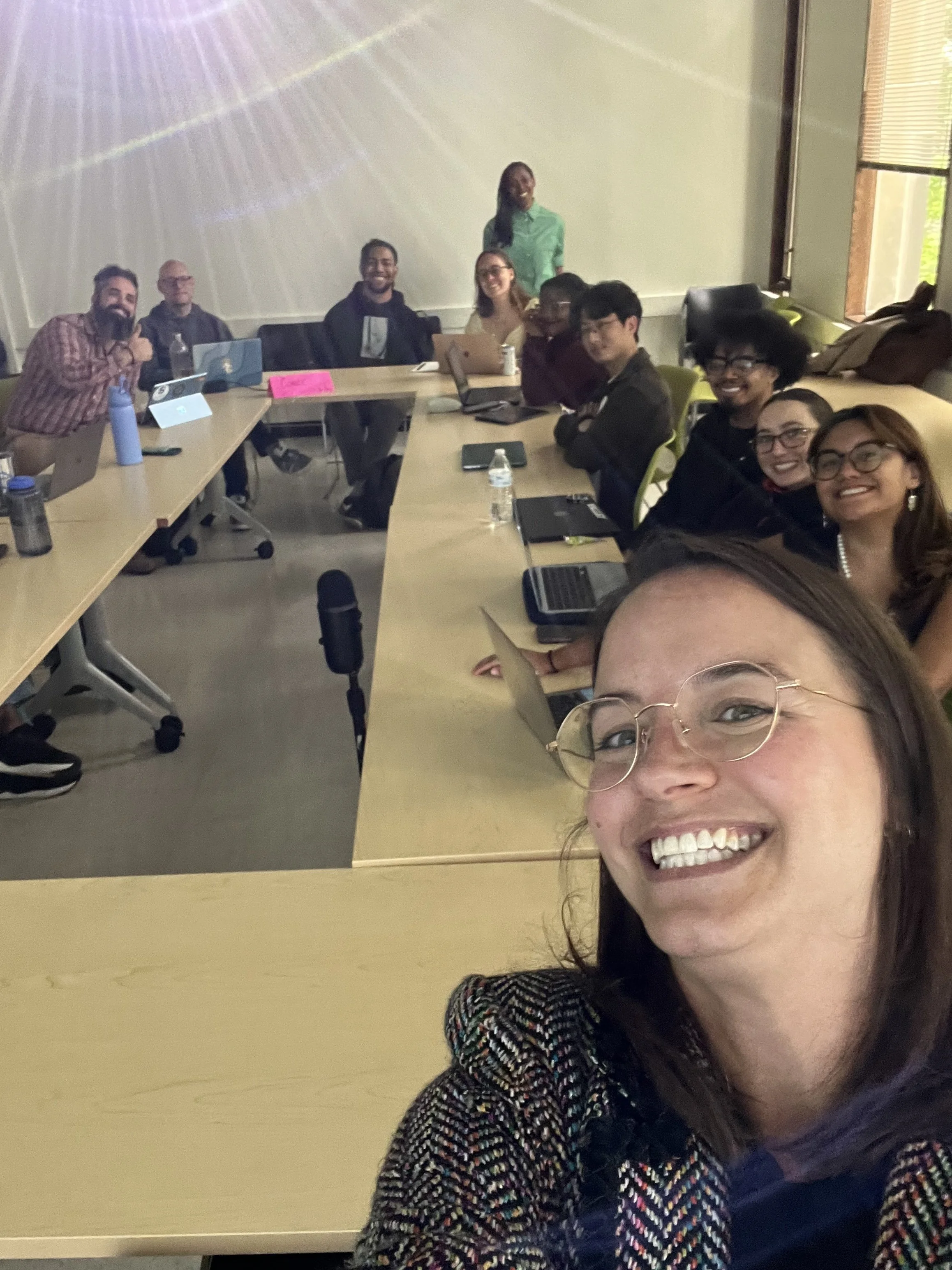 white woman with glasses taking a selfie with a classroom of students behind her