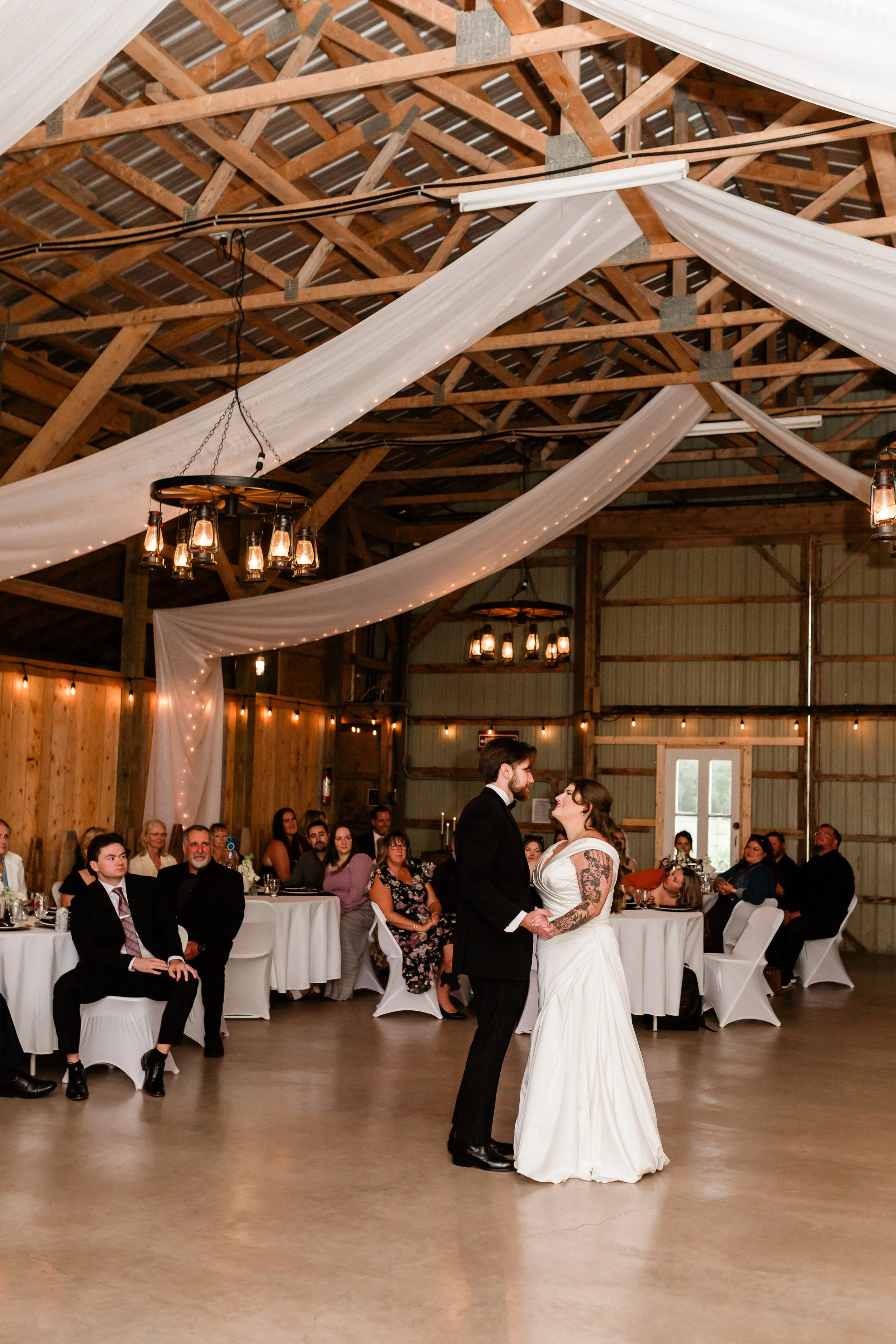 A bride and groom dance at their wedding reception in a rustic barn. The ceiling is decorated with white drapes and string lights, and guests are seated at round tables with white tablecloths.