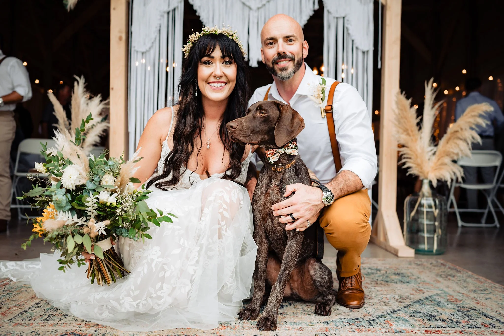 A smiling bride and groom with a brown dog at their wedding celebration indoors. The bride wears a white dress with lace details and a floral headpiece, holding a bouquet of white and green flowers. The groom is dressed in a white shirt with suspende
