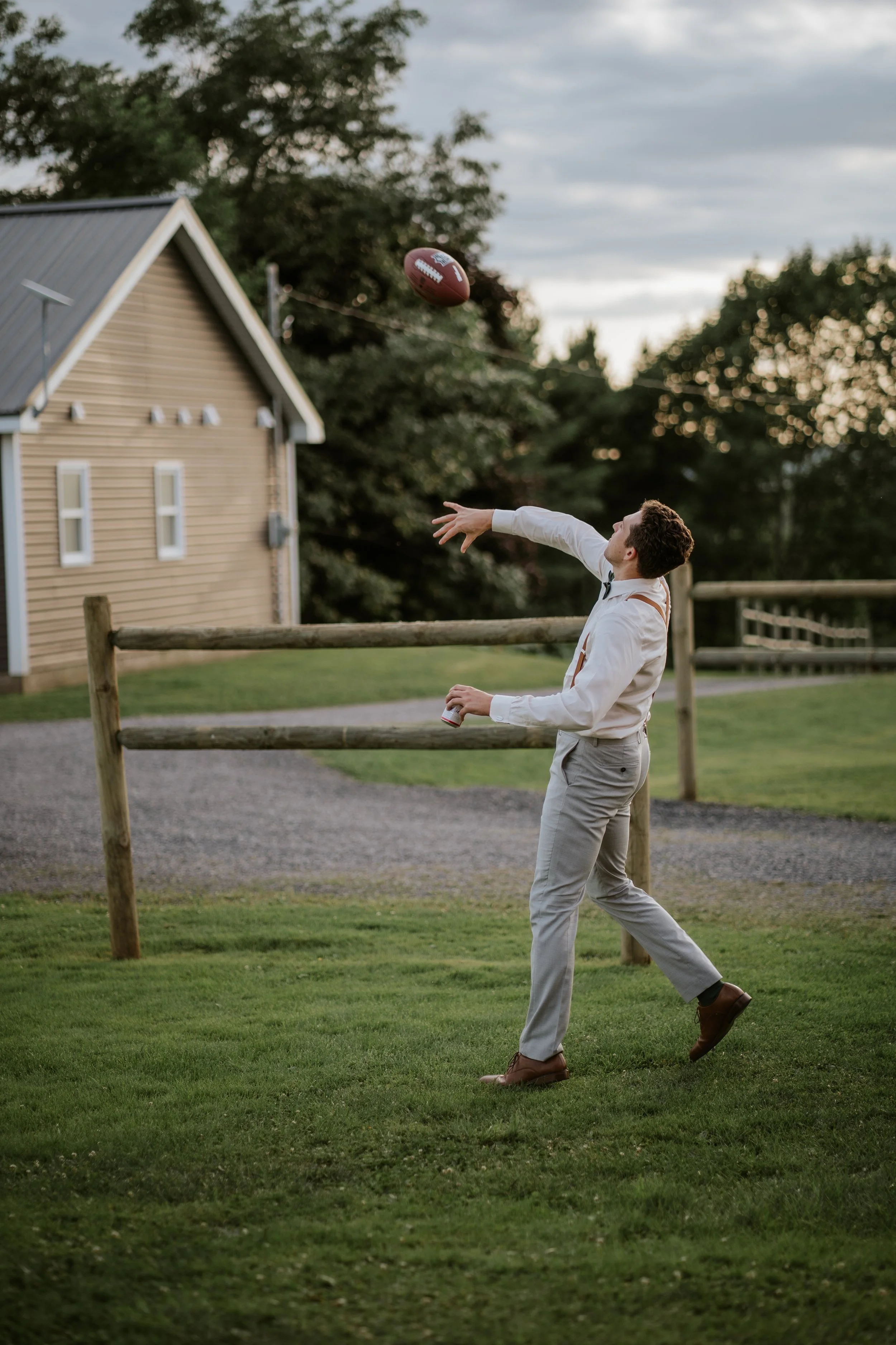 A man in dress clothes throws a football in a grassy backyard area during daylight, with a house and trees in the background.