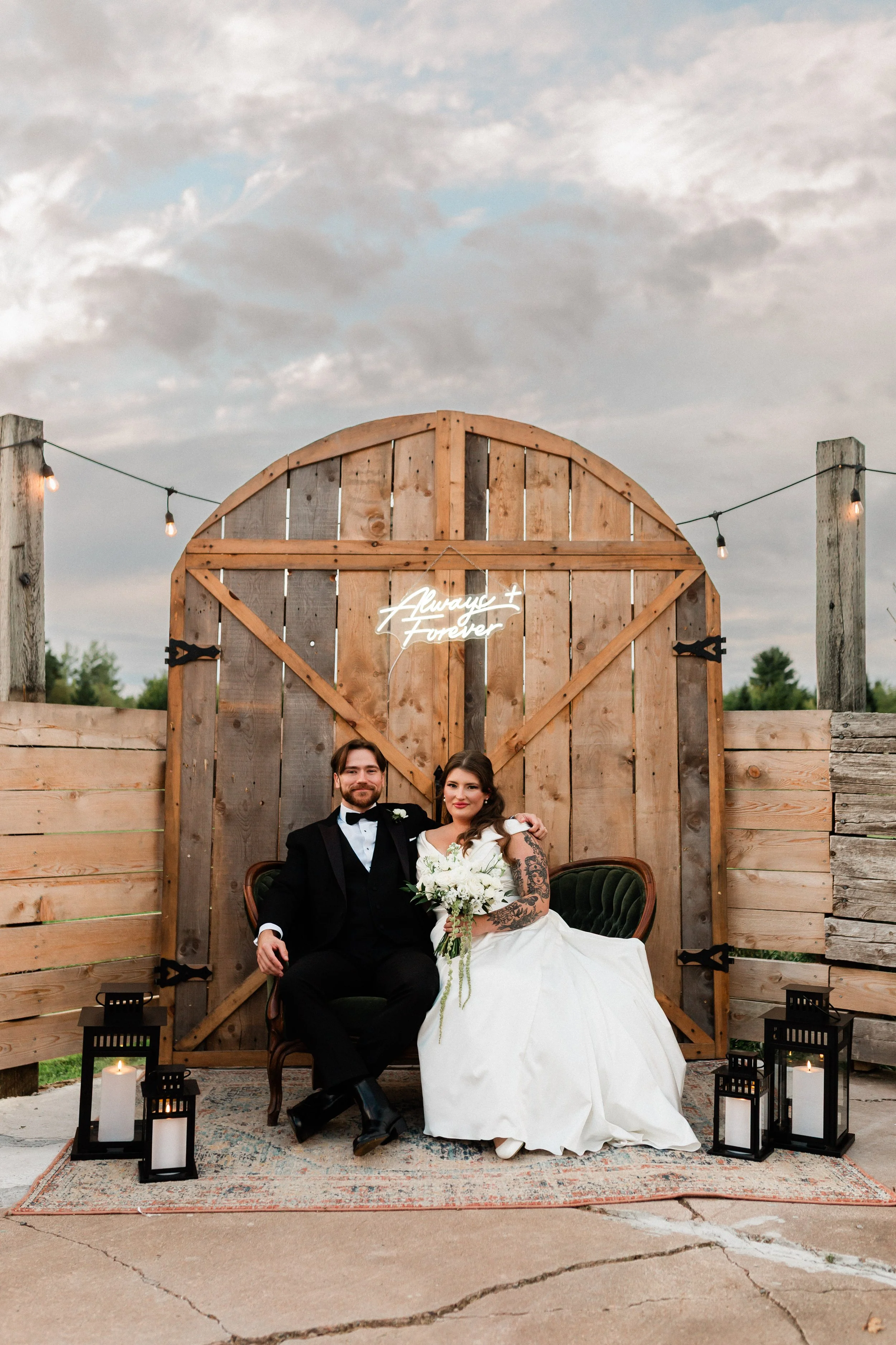 Couple in wedding attire sitting on vintage chairs outdoors, with rustic wooden backdrop, string lights, and candles, celebrating their wedding.