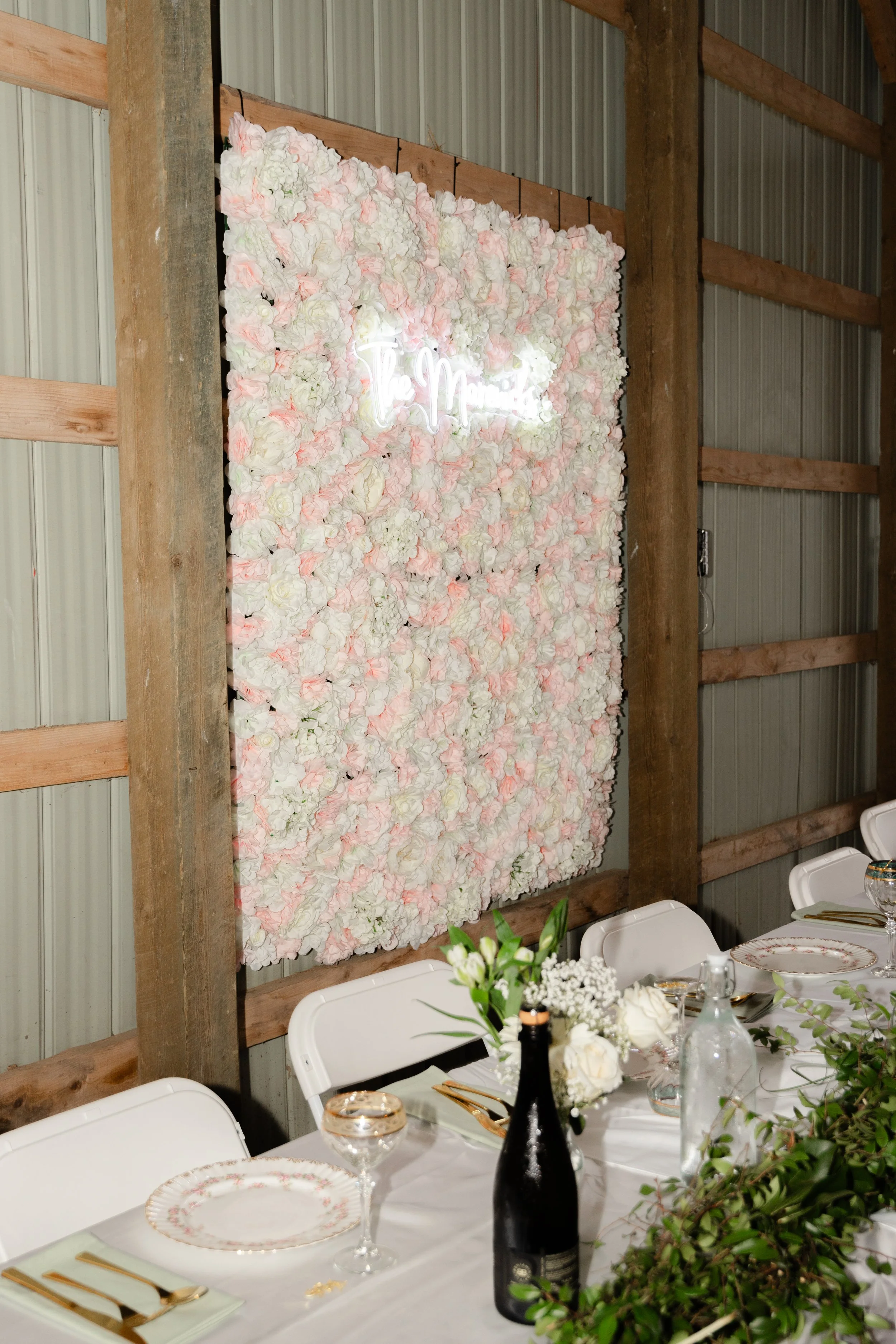 A floral wall with white and pink flowers and a neon sign that says 'The Mom.' A table with white chairs, green and white floral centerpieces, gold and white tableware, and a clear glass bottle.