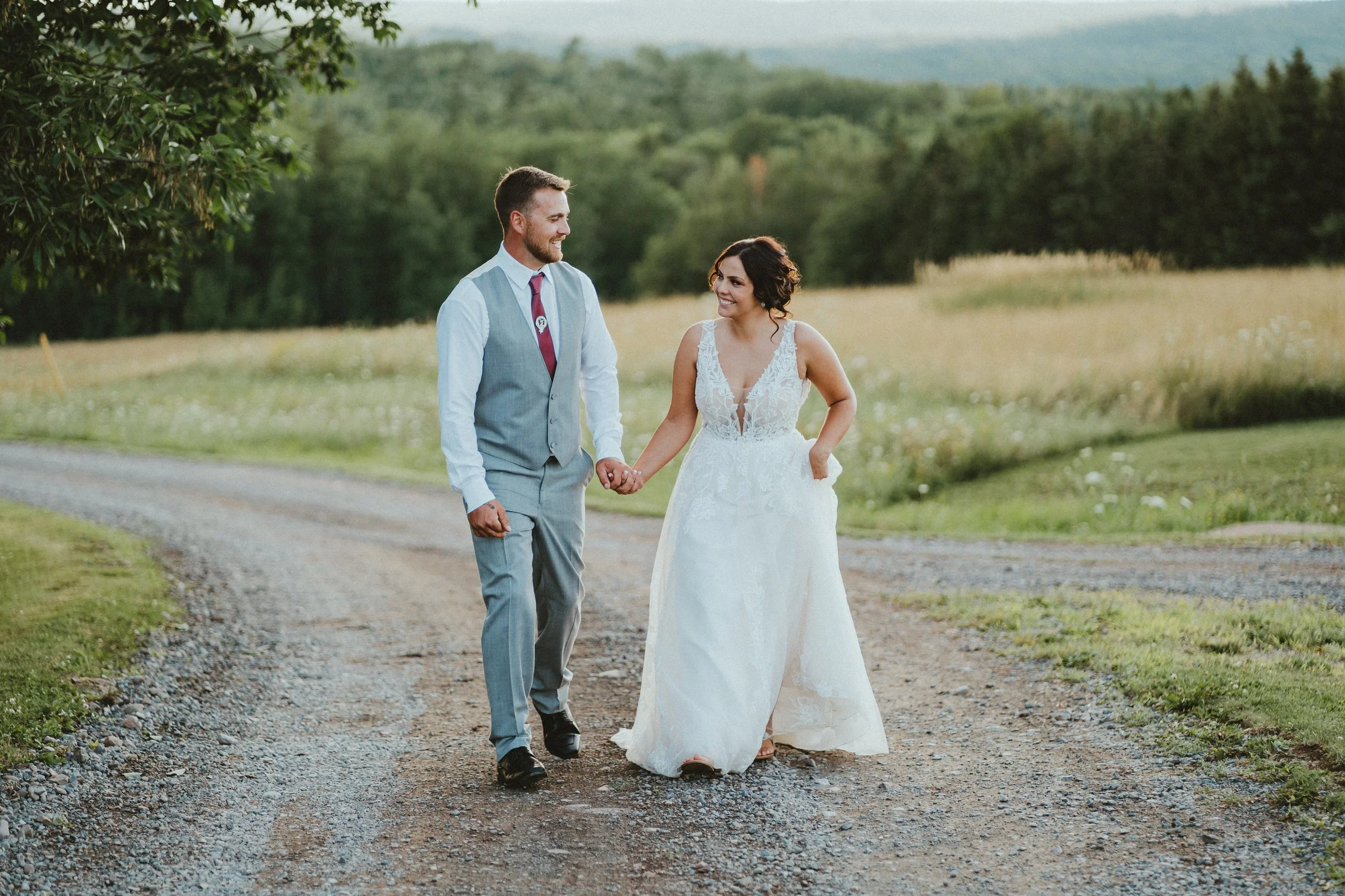A newlywed couple holding hands and walking on a gravel path in a rural landscape, with green fields and trees in the background, during sunset.