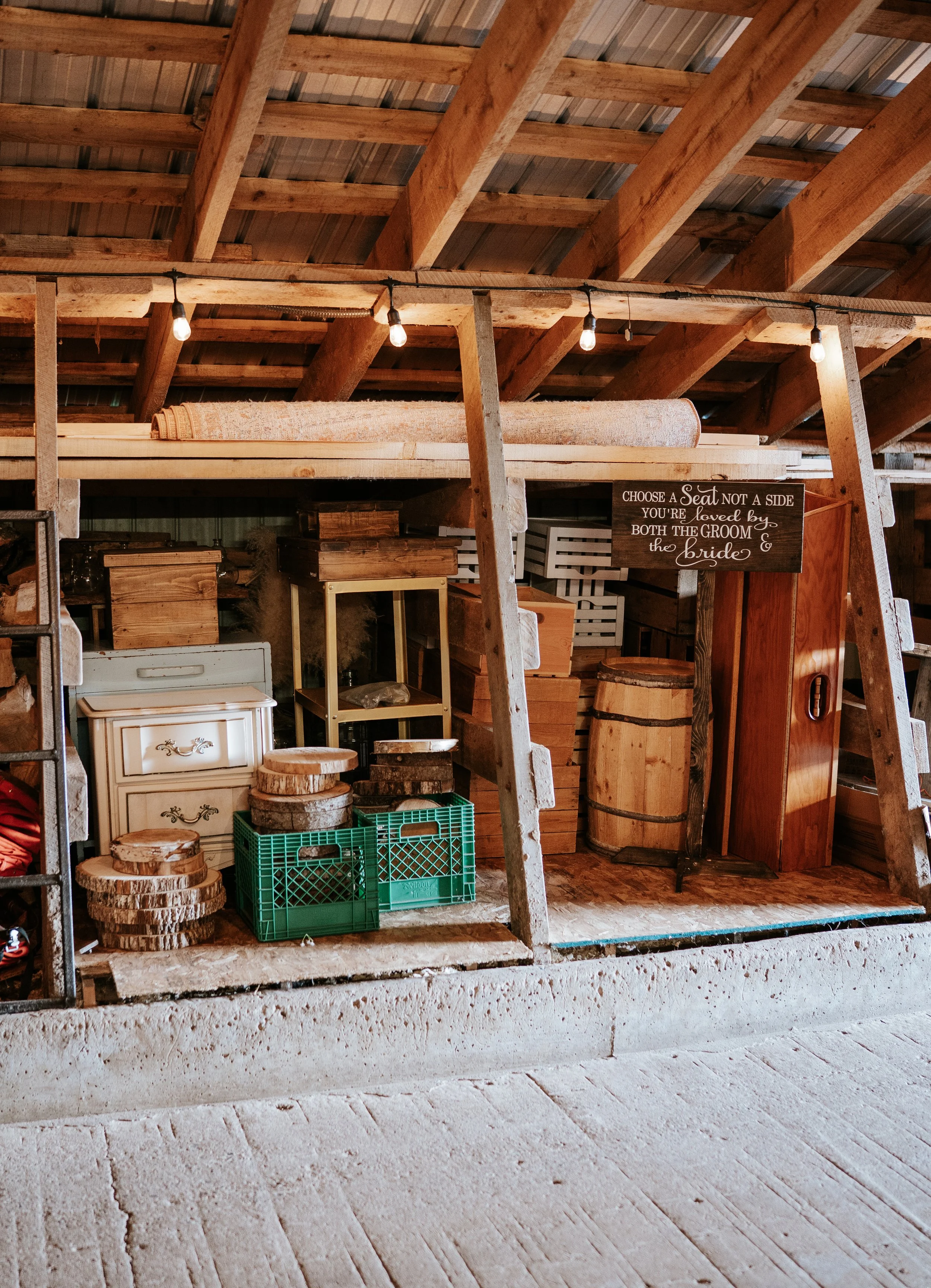 Wooden shelving unit holding various wooden rounds, crates, and furniture pieces inside a rustic barn-style building with string lights overhead.