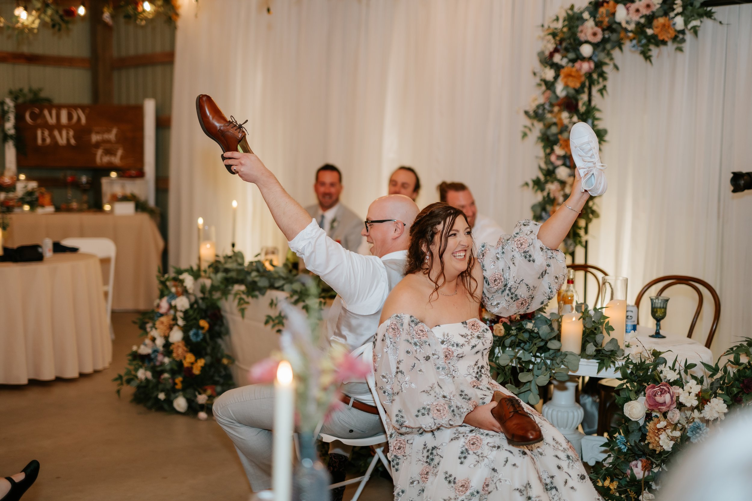 A man and woman sitting back to back, each holding a shoe in the air and smiling, at a wedding reception with guests and floral decorations.