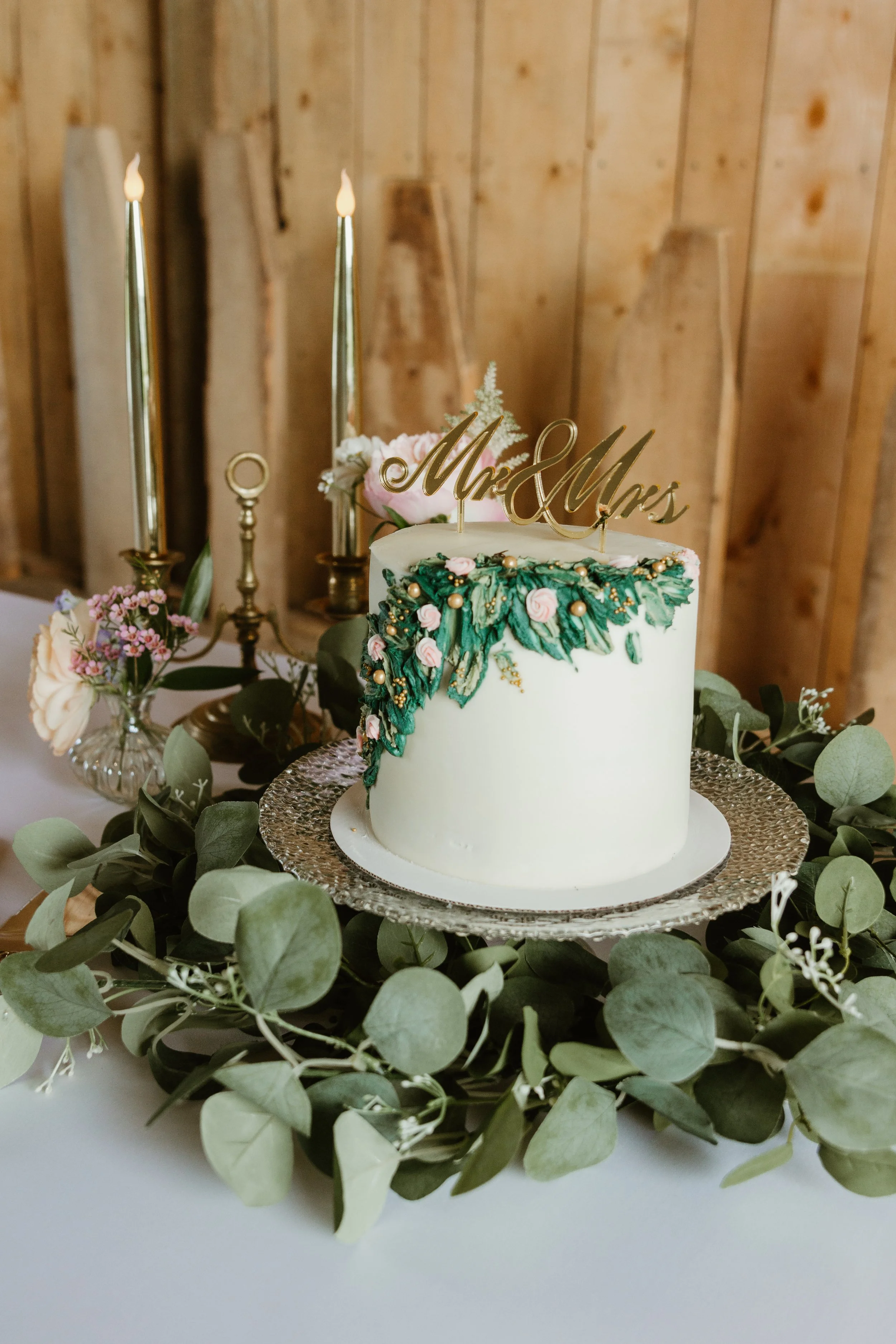 A white wedding cake decorated with green leaves, pink flowers, and gold accents, topped with a gold "Mr & Mrs" cake topper, surrounded by a green wreath of eucalyptus leaves, with gold candlesticks and pink flowers in the background.