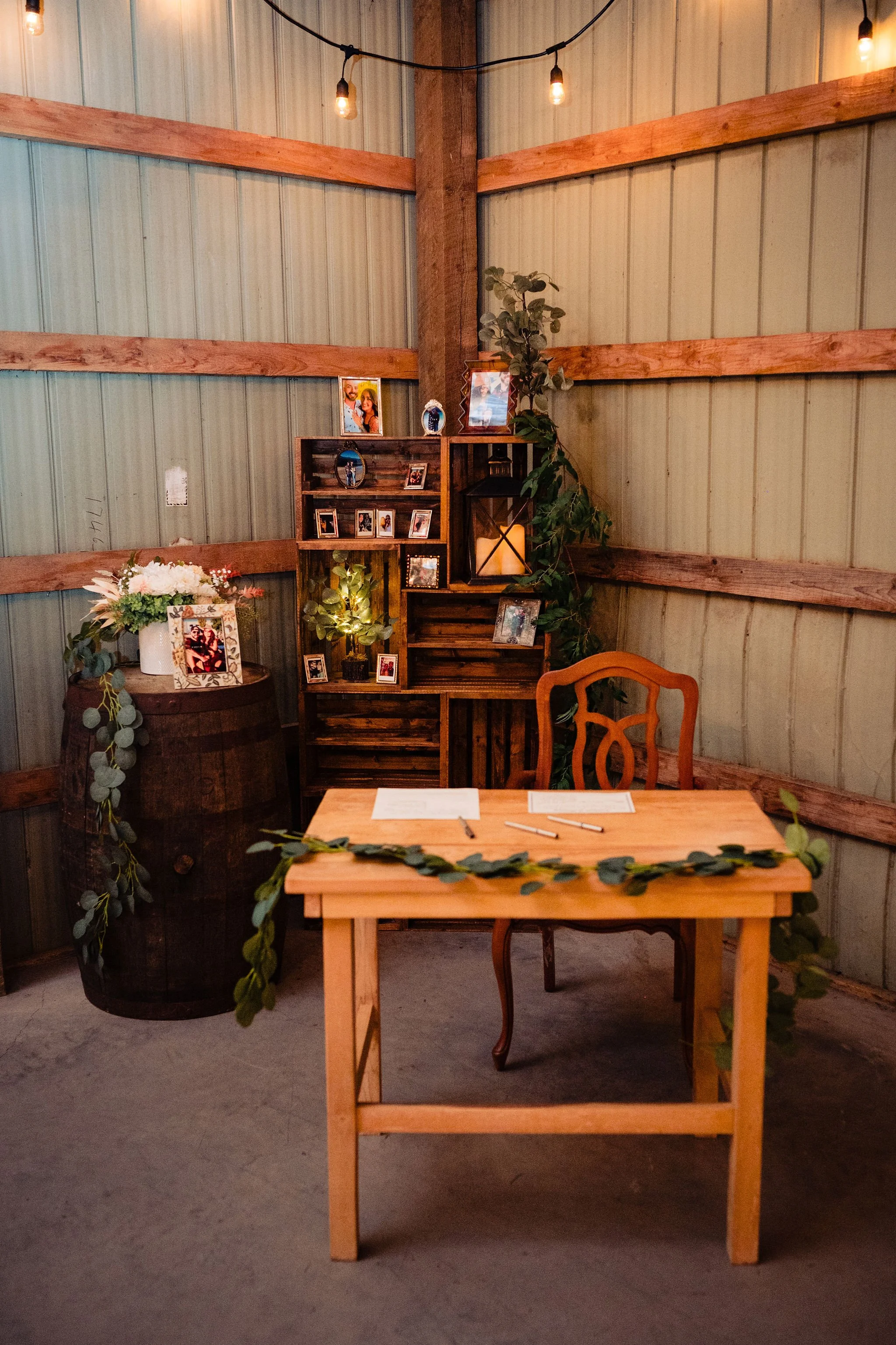 A rustic wedding or event setup with a wooden table, a vintage chair, and a decorative backdrop. The backdrop features photo frames, a lantern with a candle, greenery, and string lights hanging from the ceiling. A flower arrangement is placed on a wo