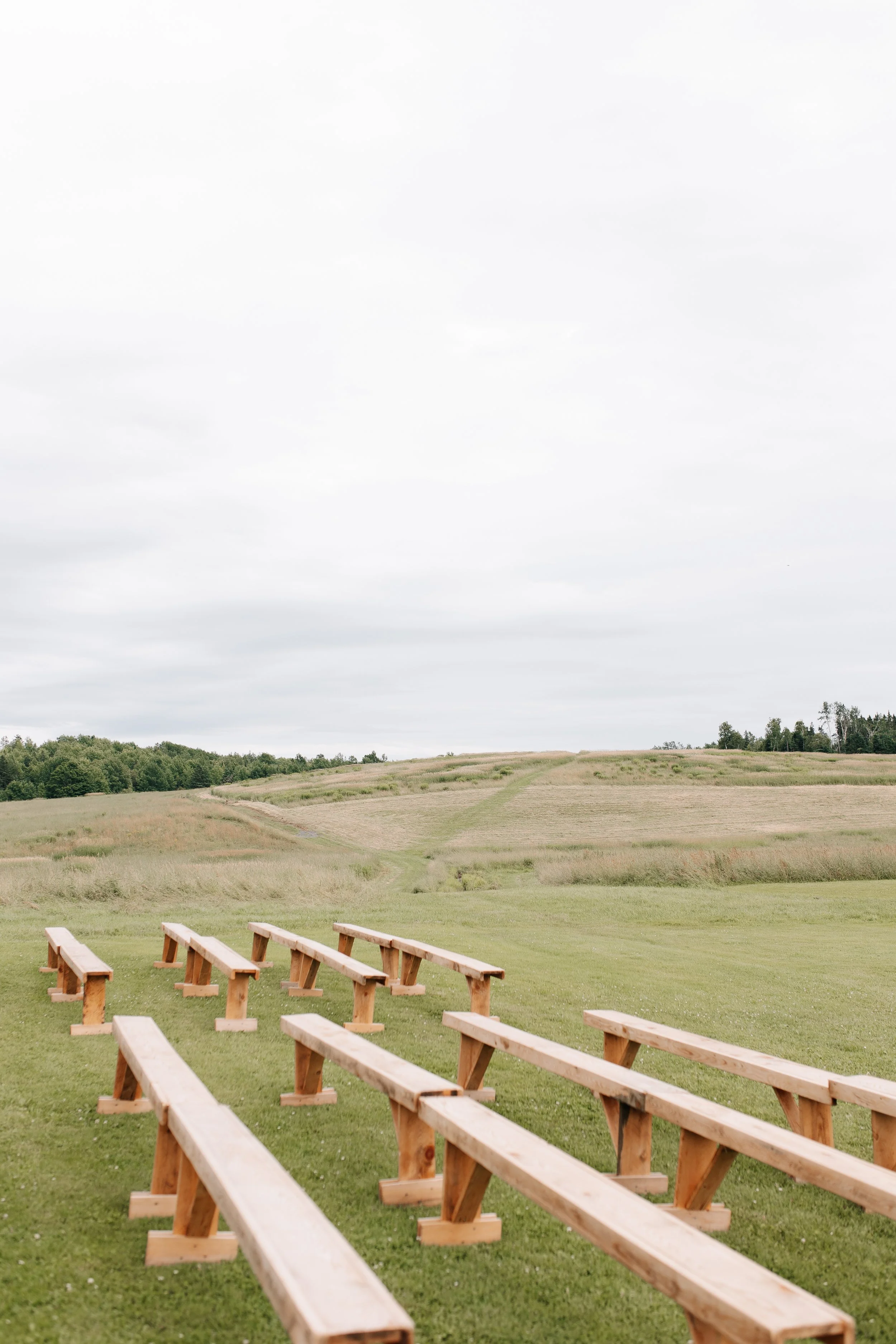 An outdoor scene with wooden benches arranged on green grass, facing a distant rolling hill under a cloudy sky.