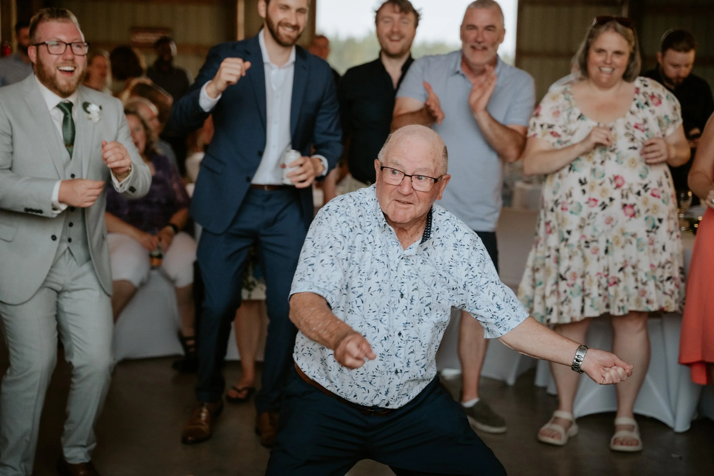 An elderly man is dancing at a celebration, surrounded by smiling guests, some of whom are clapping and cheering.