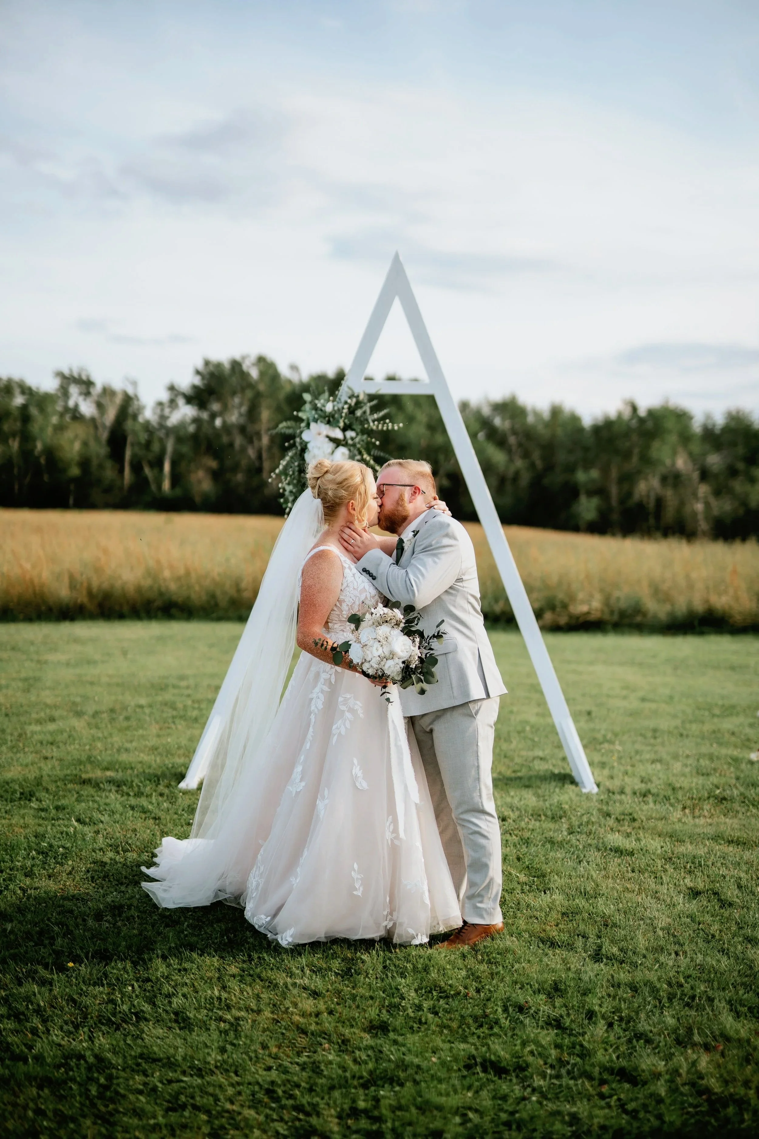 A bride and groom share a kiss during their outdoor wedding ceremony, standing in front of a white triangular arch decorated with flowers and greenery, on a grassy field with trees in the background.