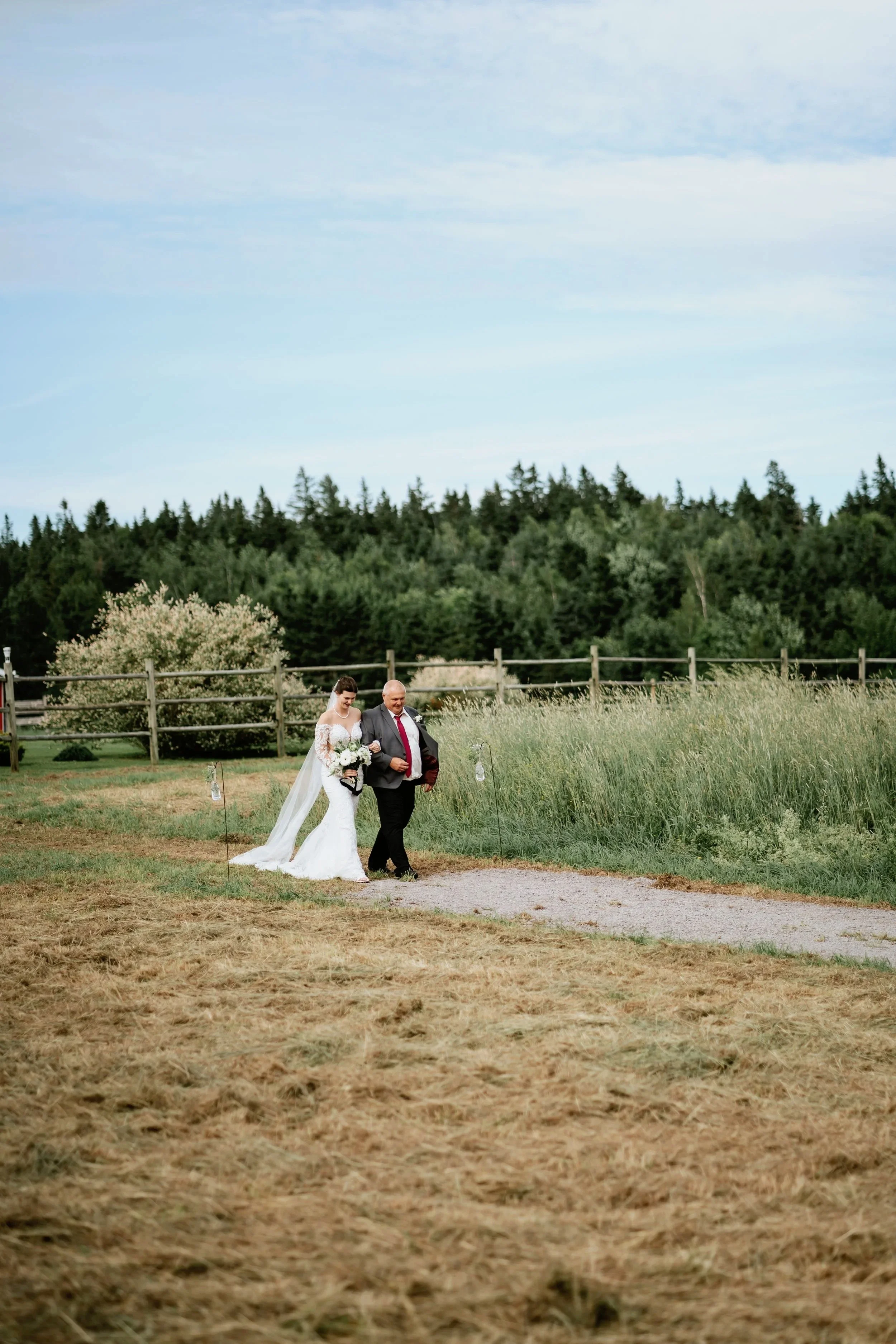 A bride and a man, possibly her father, walking outdoors on a gravel path during a wedding. The bride is in a white wedding gown with a long veil, holding a bouquet of flowers. The man is in a dark suit with a red tie. The background includes green t