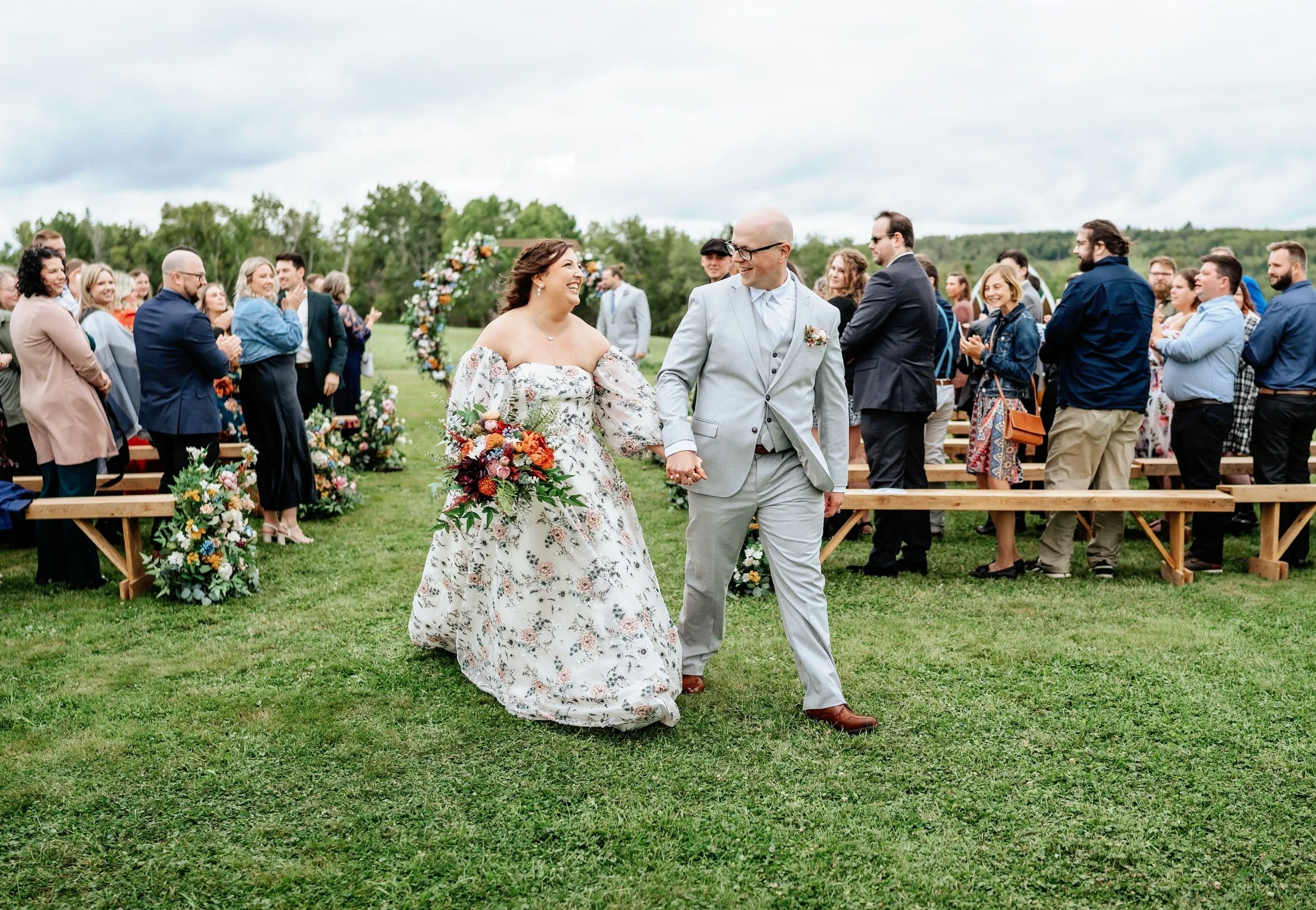 A bride and groom walking hand in hand down the aisle at an outdoor wedding ceremony, smiling and looking at each other, with guests clapping and celebrating in the background, overcast sky with trees in the distance.