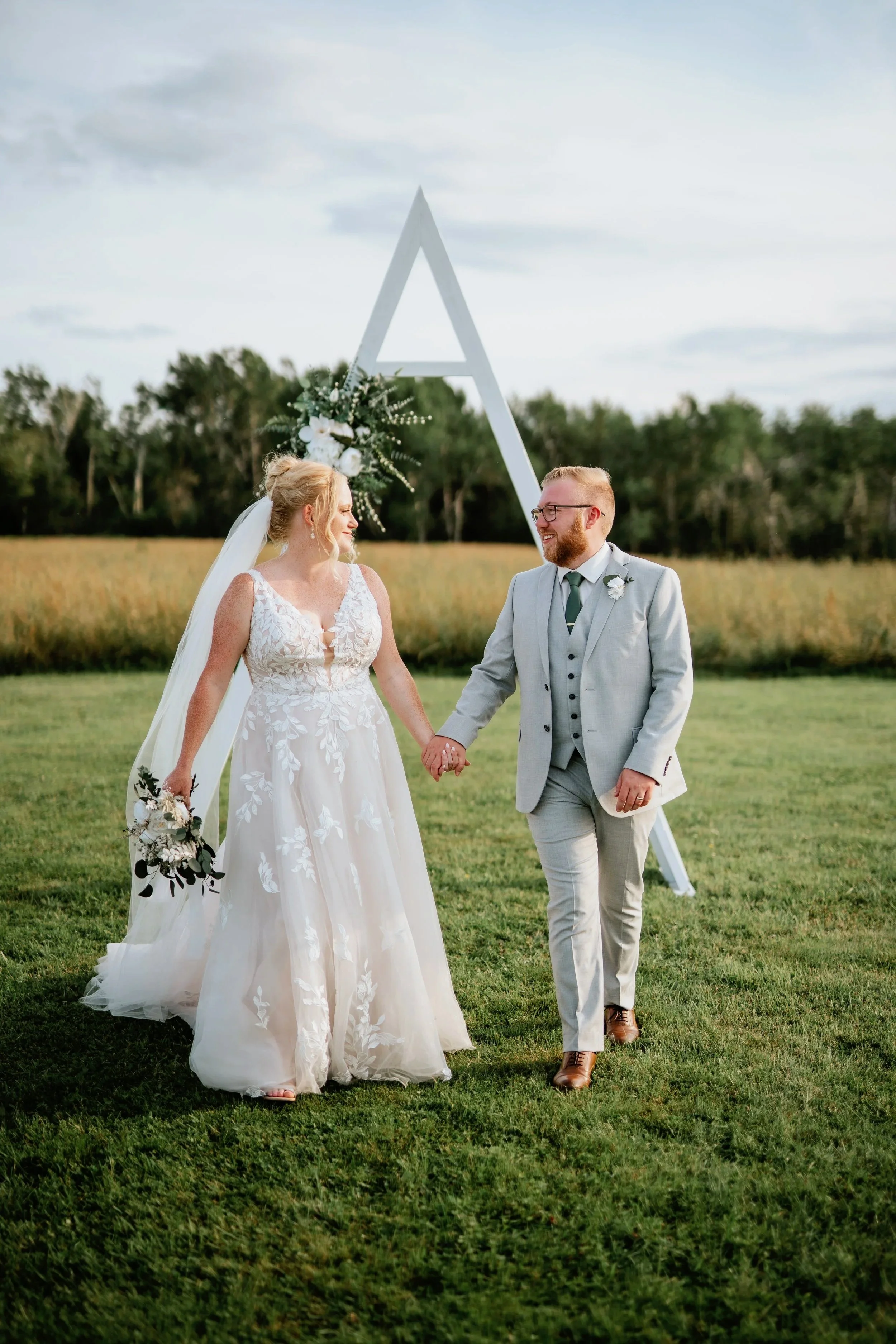 A bride and groom holding hands and walking on a grassy field during their outdoor wedding, with a large white triangular decoration and greenery in the background.