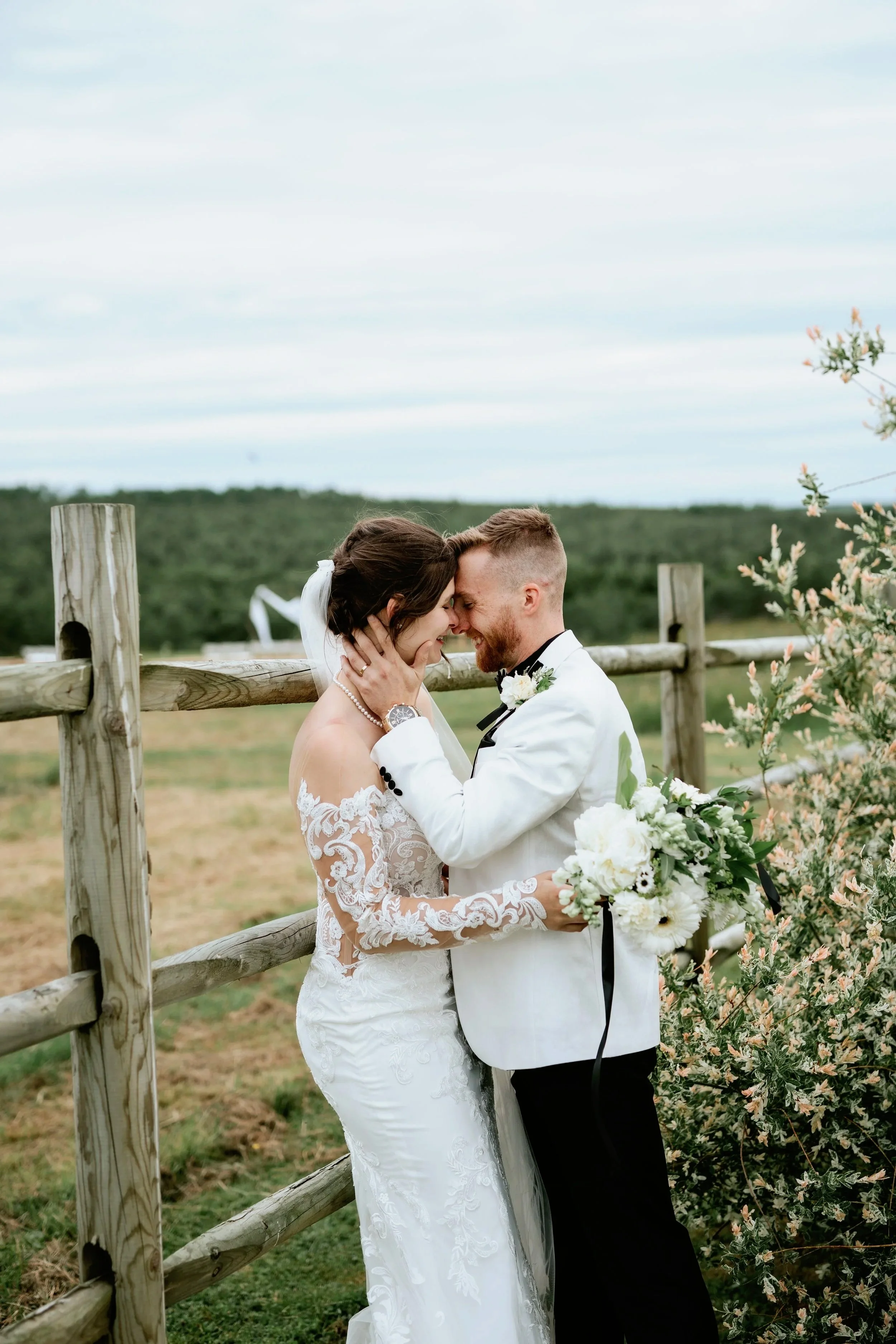 A bride and groom sharing a joyful moment outdoors, with the bride in a lace wedding dress holding a bouquet, and the groom in a white tuxedo jacket, touching foreheads with their eyes closed.