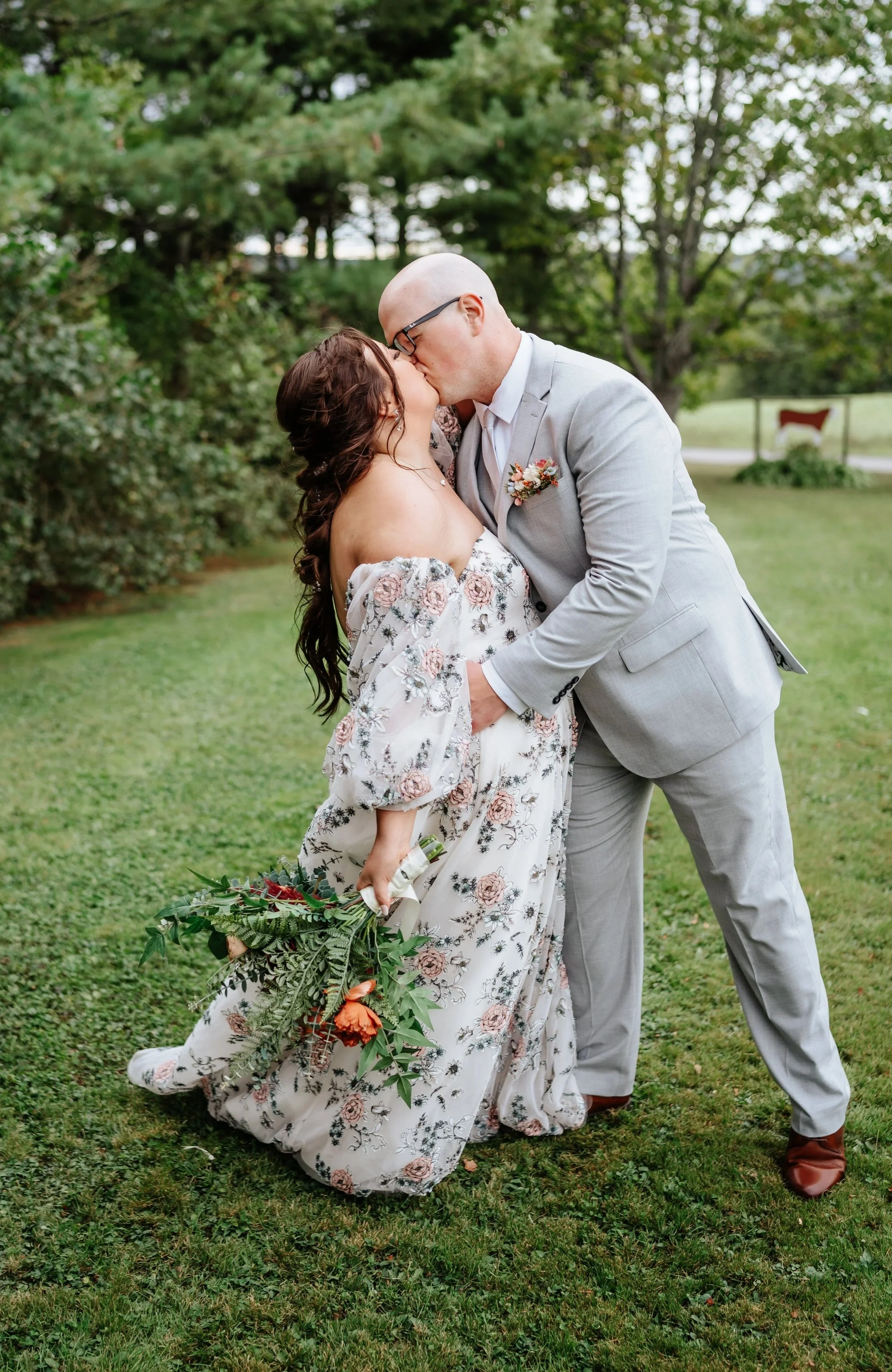 A newlywed couple sharing a kiss outdoors on a grass lawn, with the bride holding a bouquet of flowers and wearing a floral off-the-shoulder gown, and the groom in a light gray suit with a boutonniere.