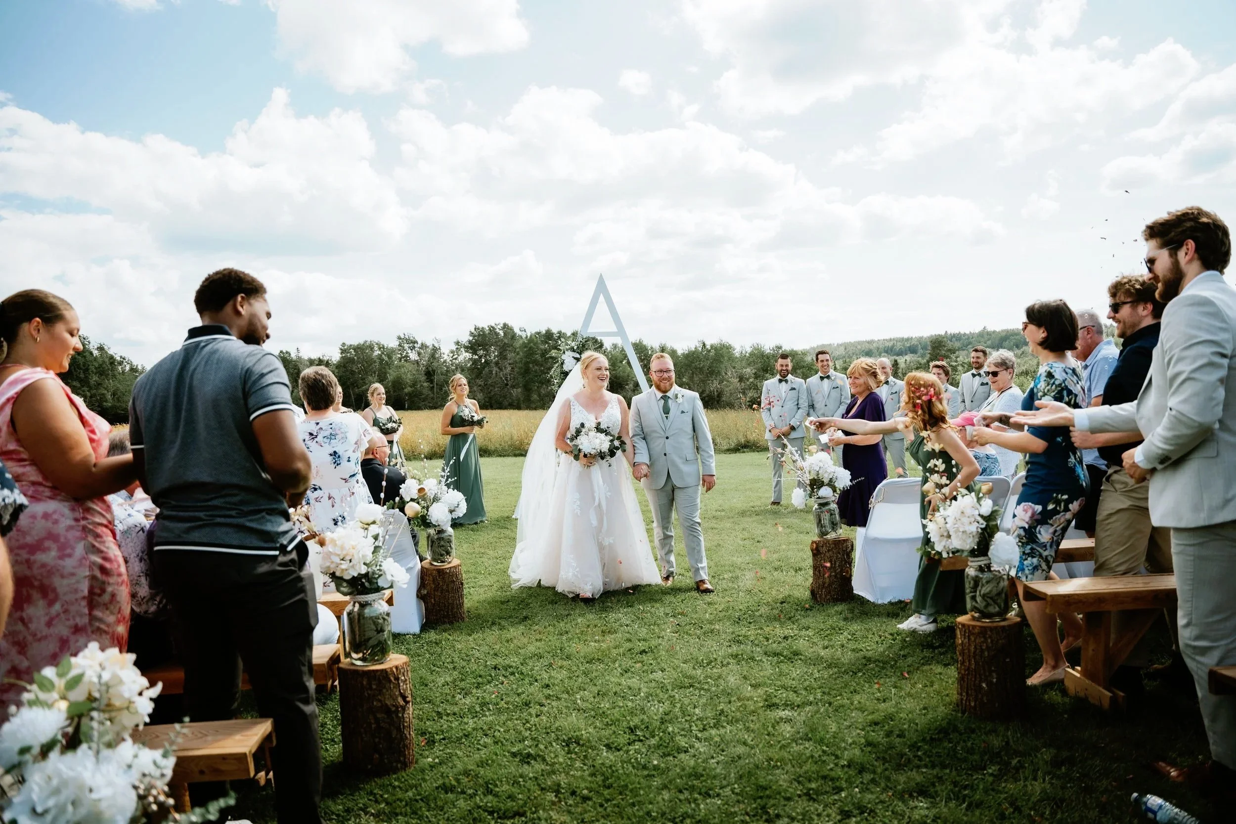 Wedding ceremony outdoors with bride and groom walking down the aisle, surrounded by guests in casual and semi-formal attire, on a grassy field with trees and a cloudy sky background.