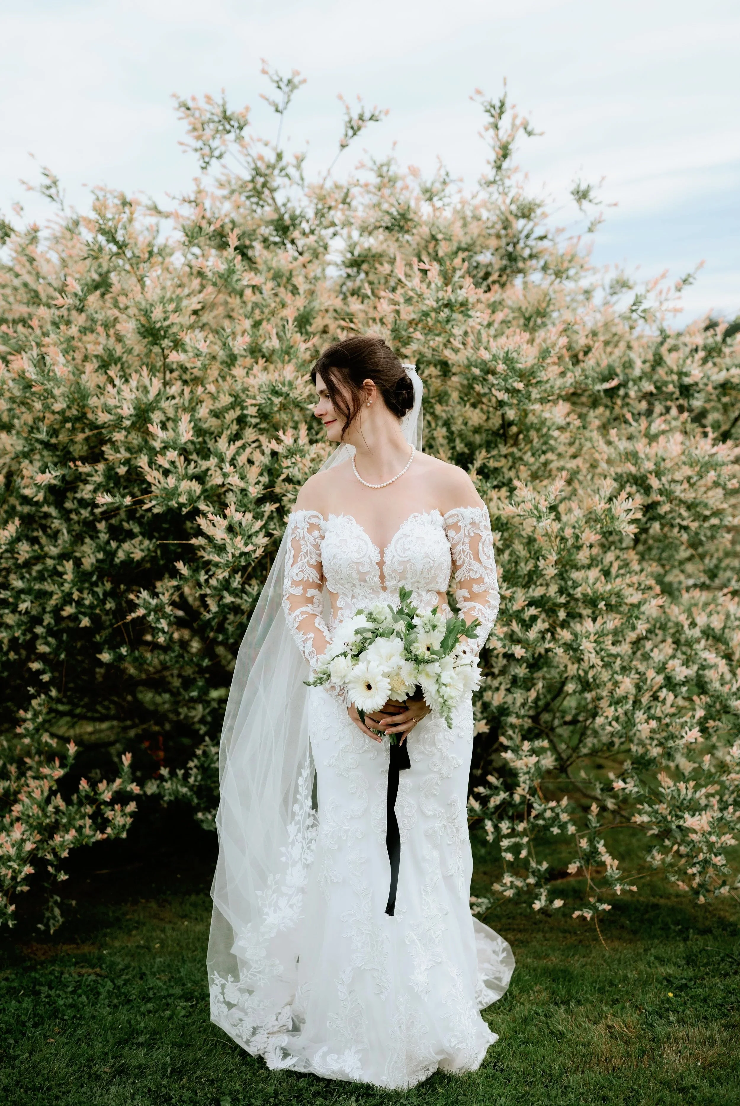 Bride in a white lace wedding dress with long sleeves, standing outdoors in front of blooming bushes, holding a bouquet of white flowers, looking to her side.