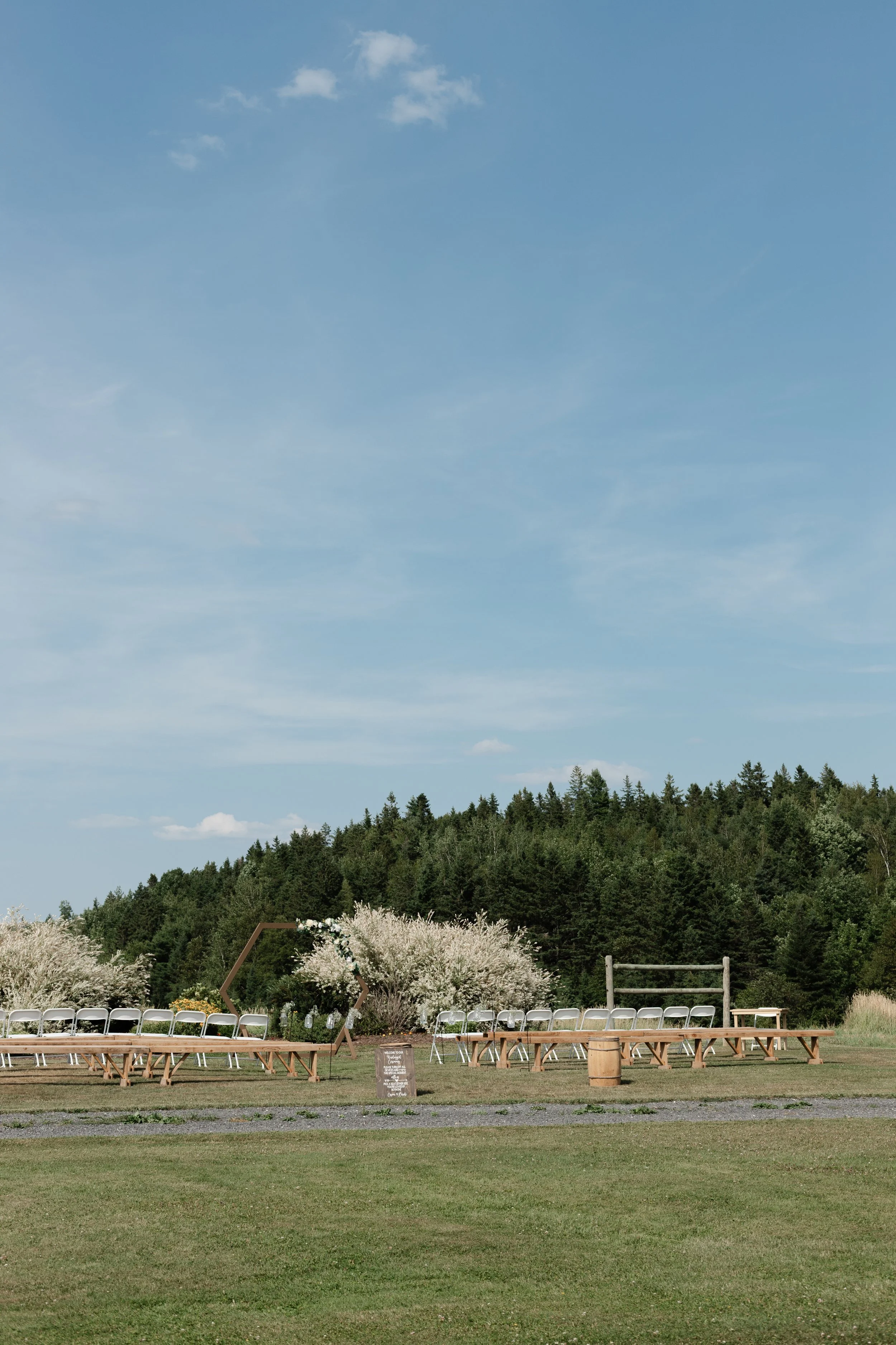Outdoor wedding ceremony setup with white chairs, a decorated hexagon-shaped arbor, and a grassy field with trees and a blue sky in the background.