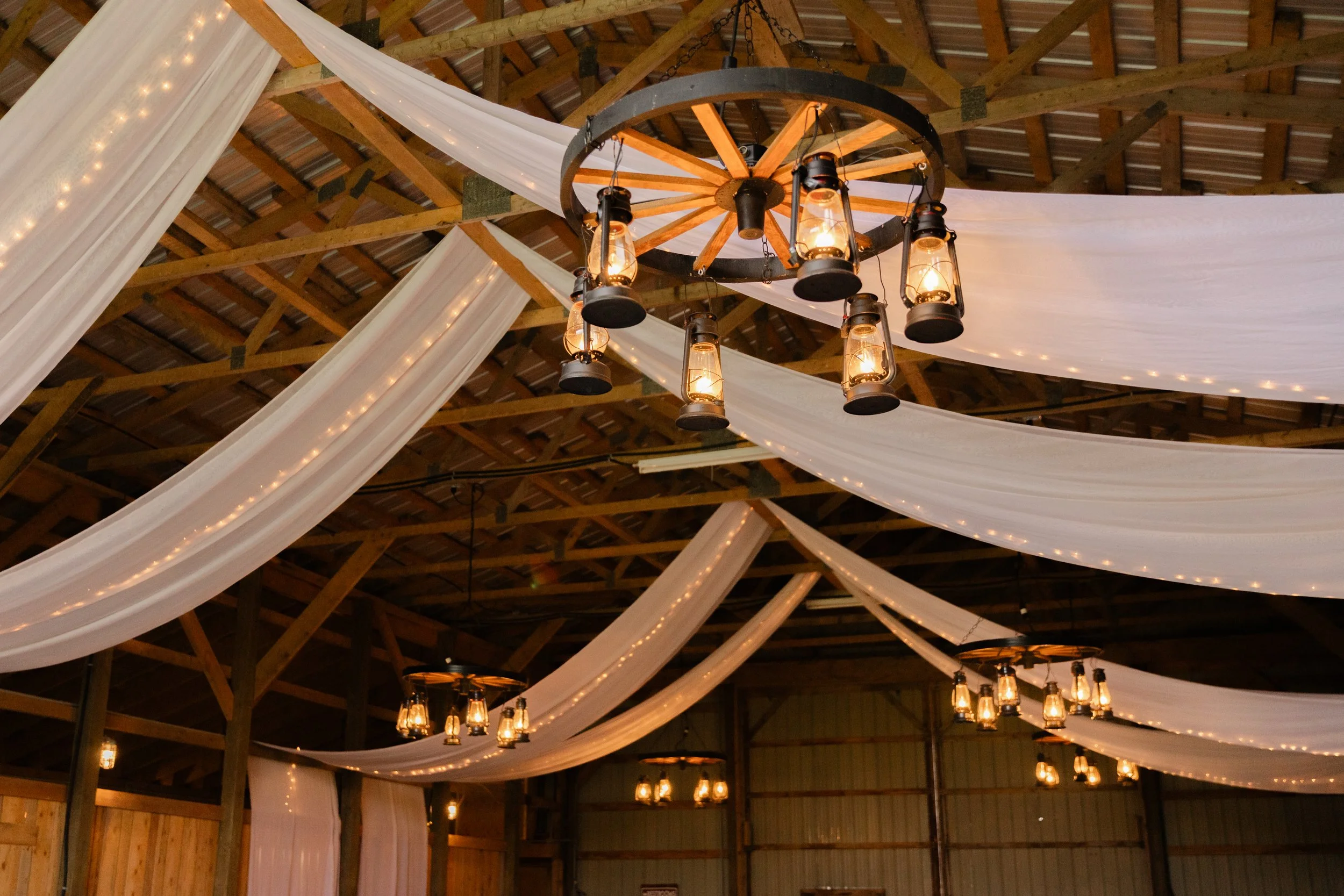 Decorative lighting with lanterns hanging from a wagon wheel chandelier, draped white fabric, and string lights inside a barn