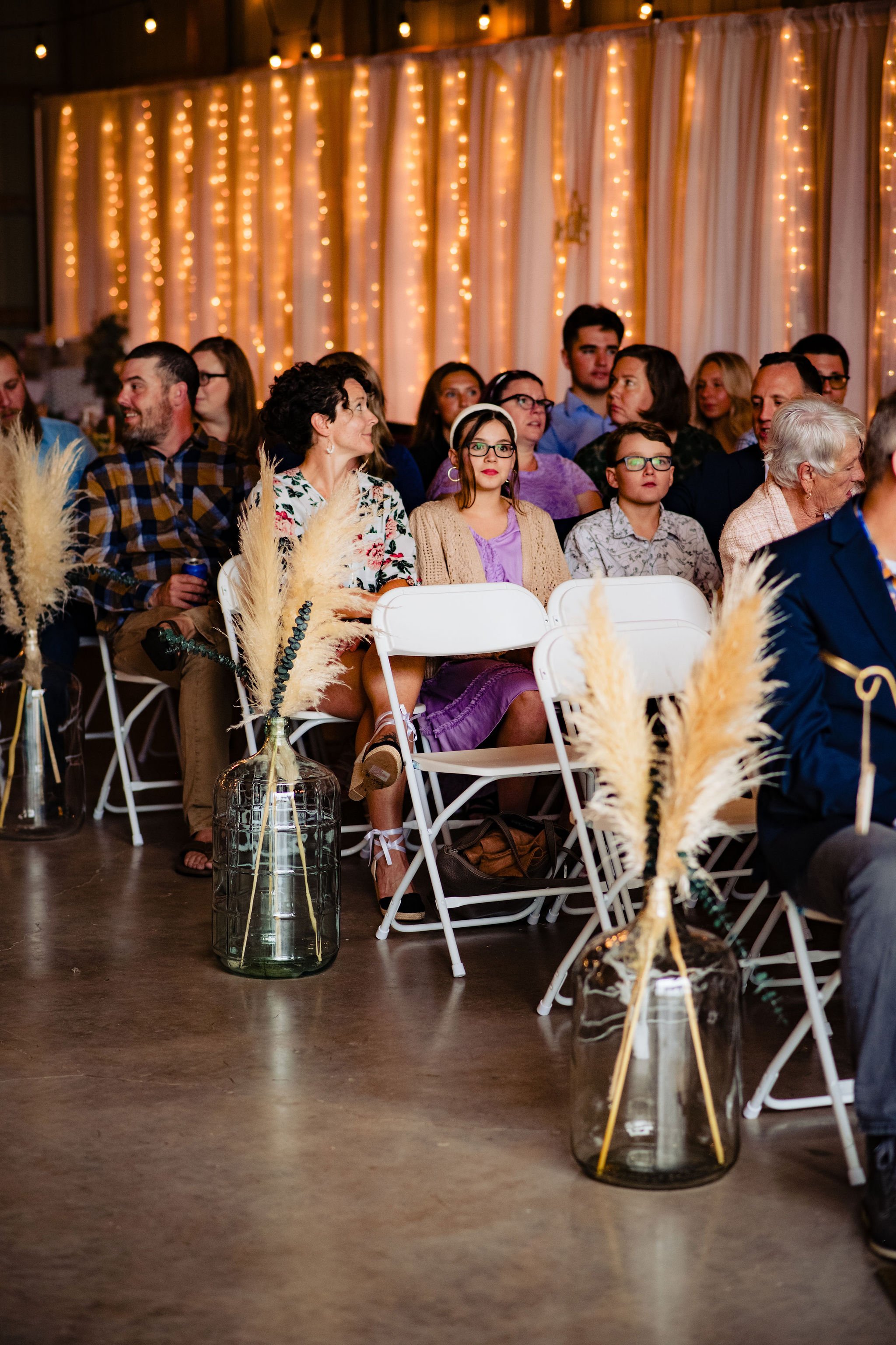 Audience members sitting at a decorated event with string lights and tall vases with pampas grass in white chairs.