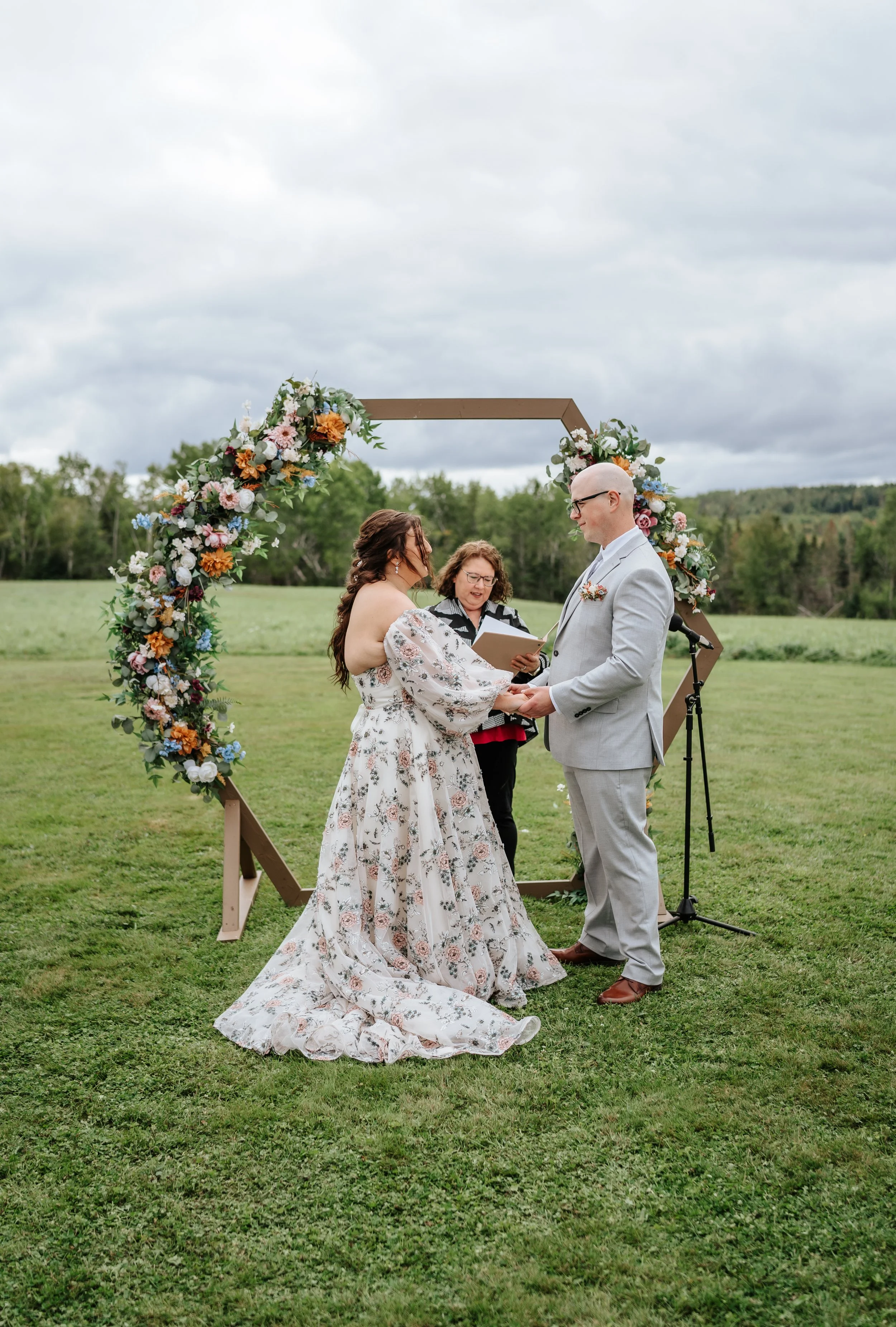 A couple getting married outdoors under a floral arch with an officiant. The bride is wearing a floral off-the-shoulder wedding gown, and the groom is in a light grey suit. They are holding hands and exchanging vows, with a scenic green field and clo