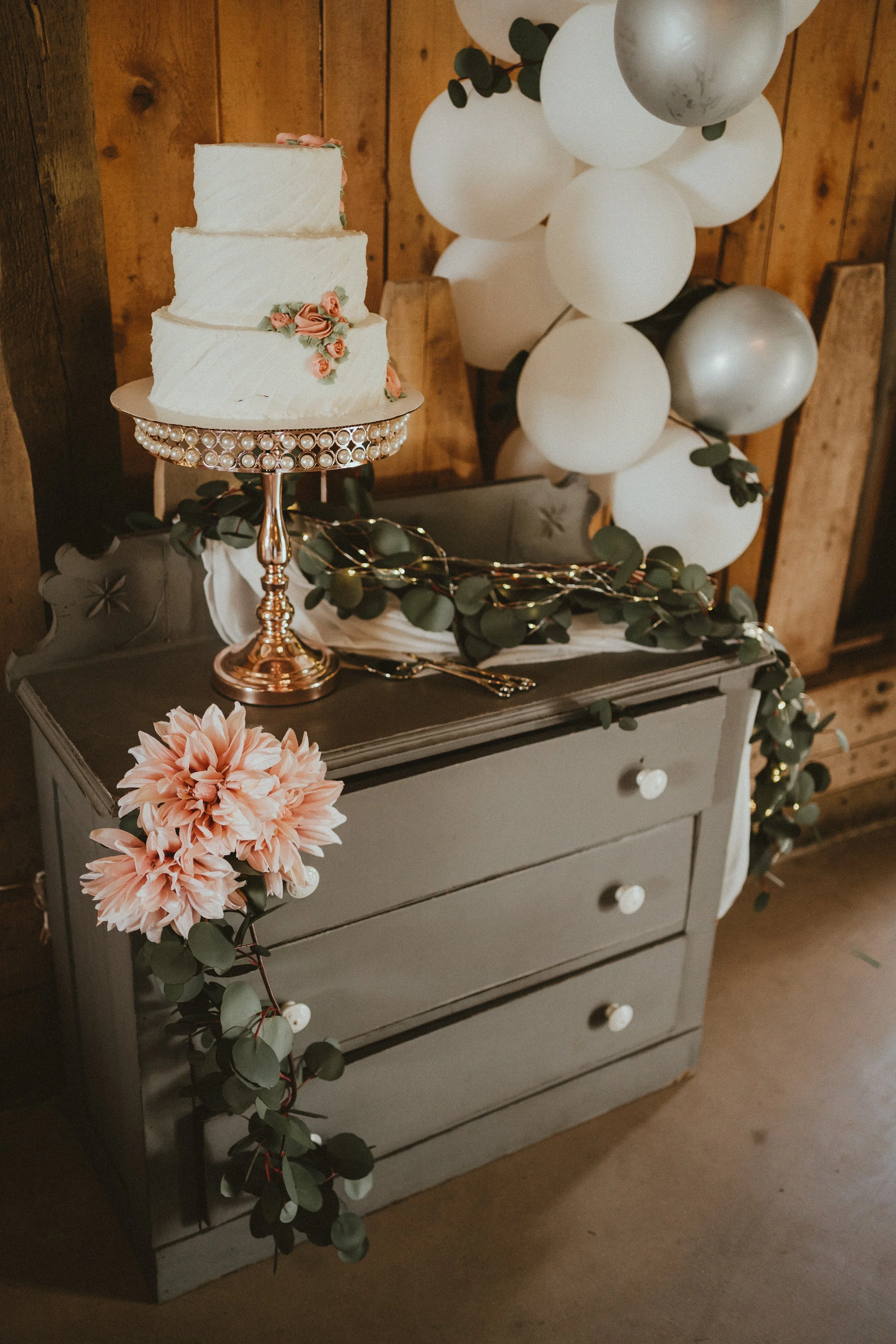 A three-tier white wedding cake with pink flowers on top, displayed on a decorative stand on a gray dresser. The dresser is decorated with a eucalyptus garland and pink flowers. White and silver balloons are in the background, with a wooden wall behi
