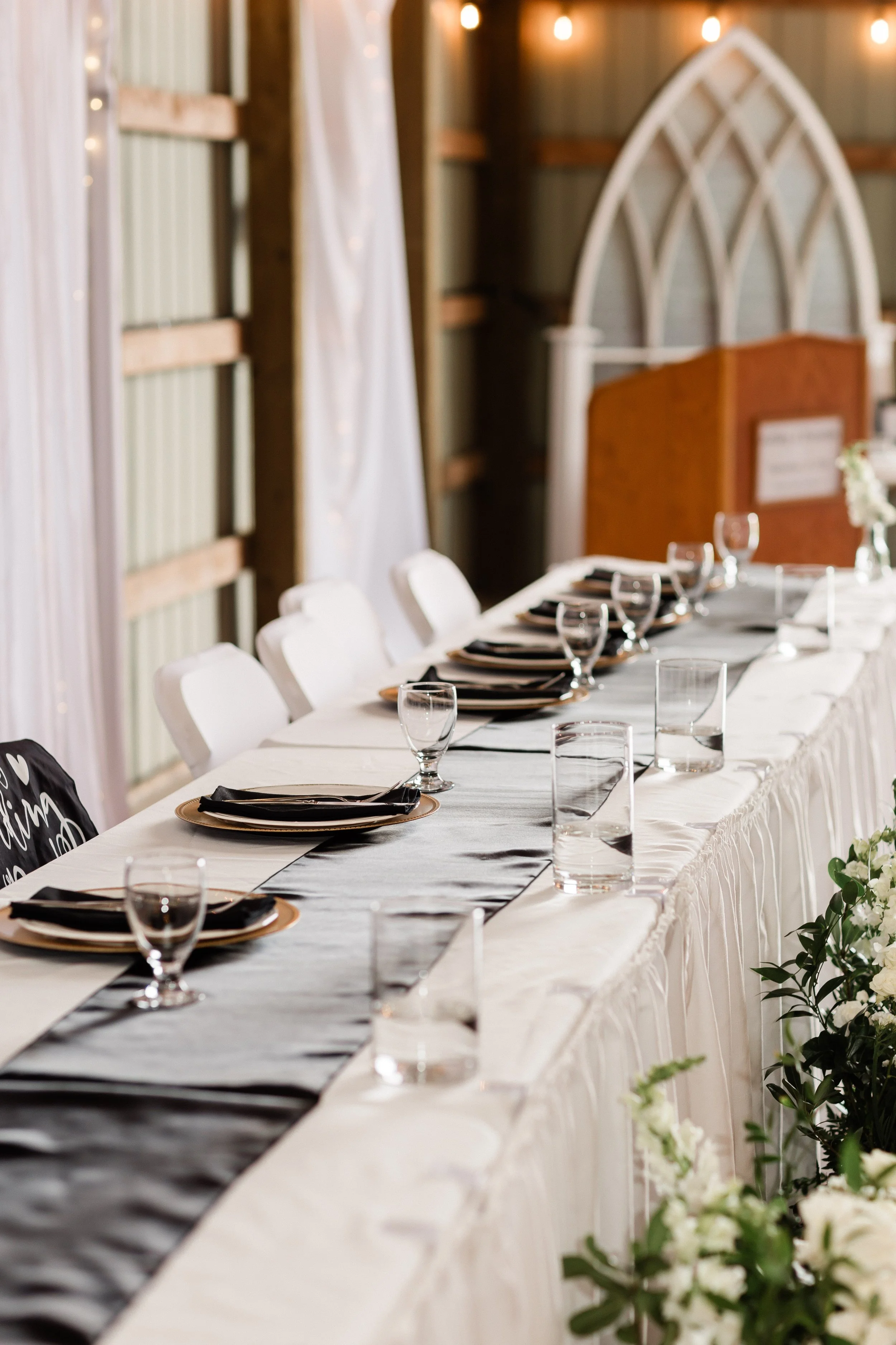 A long banquet table set for a formal event with white tablecloth, black inlay, and black napkins, complemented by plates, water glasses, and wine glasses, with floral arrangements at the end, in a rustic barn setting with string lights and wooden wa