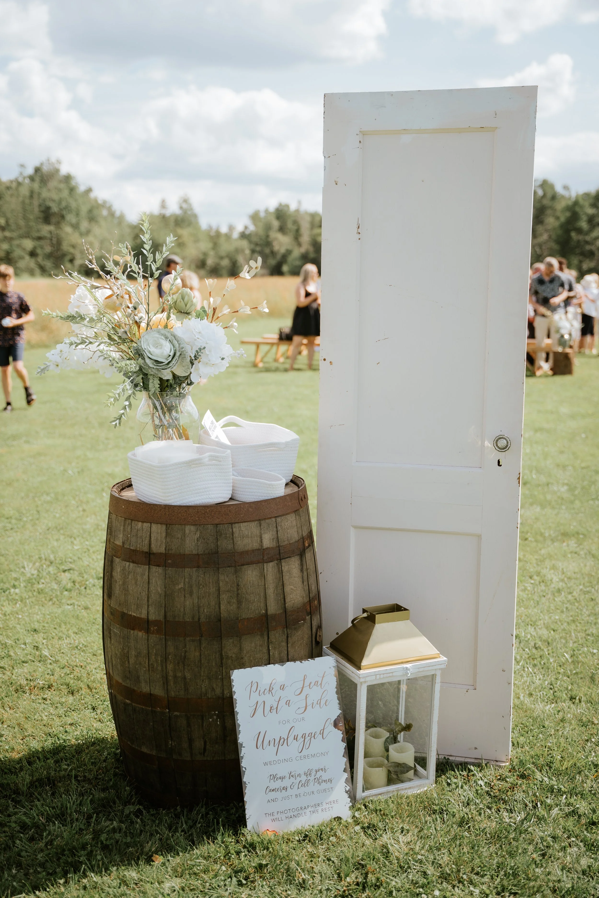 Wedding seating area with a white door, a wooden barrel with a flower arrangement, a lantern with candles, and a sign that reads "Pick a Seat Not a Side for our Unplugged Wedding Ceremony" on a grassy field with people in the background.
