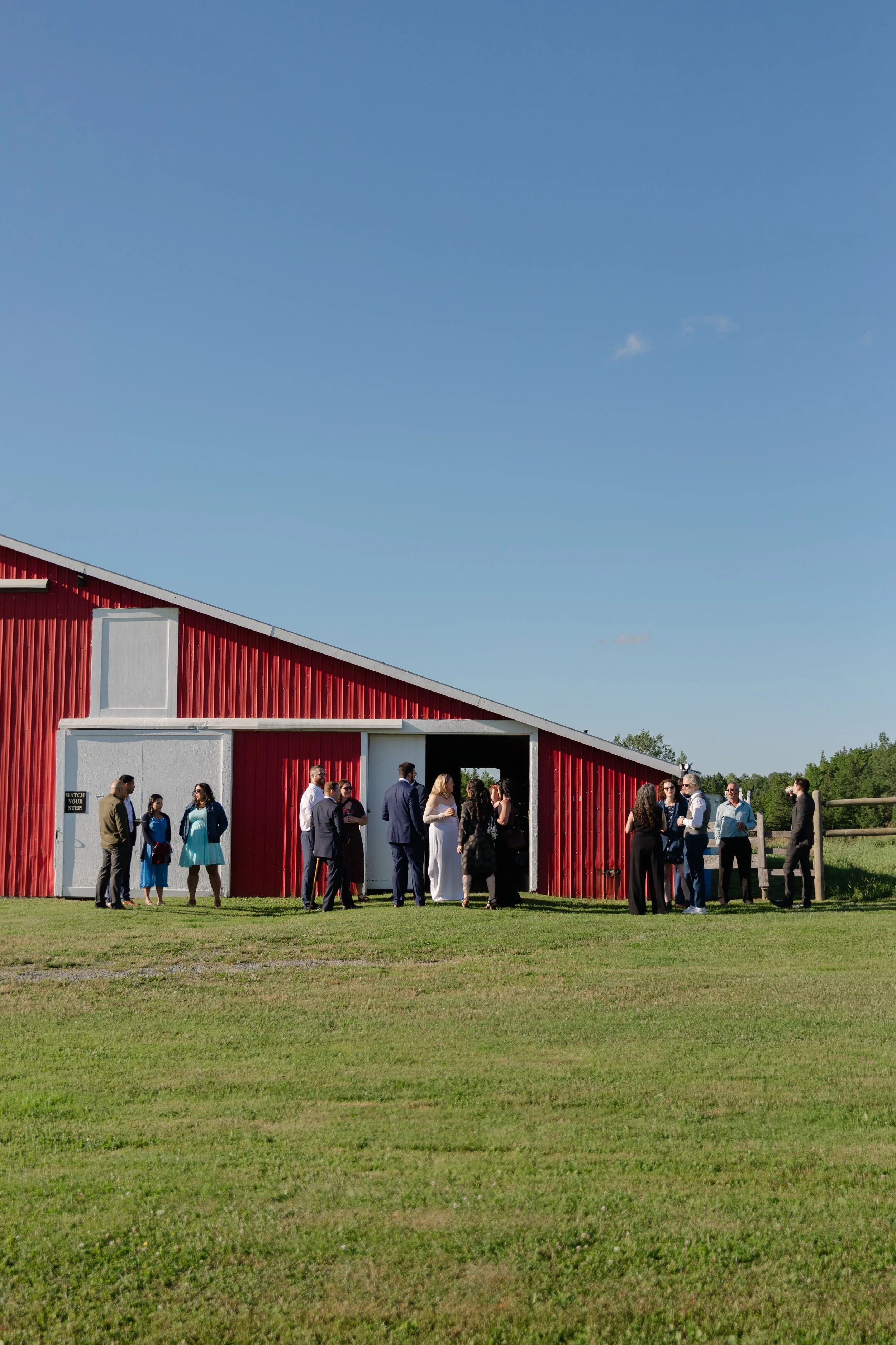 Group of people gathered outside a red barn-like building on a clear, sunny day.